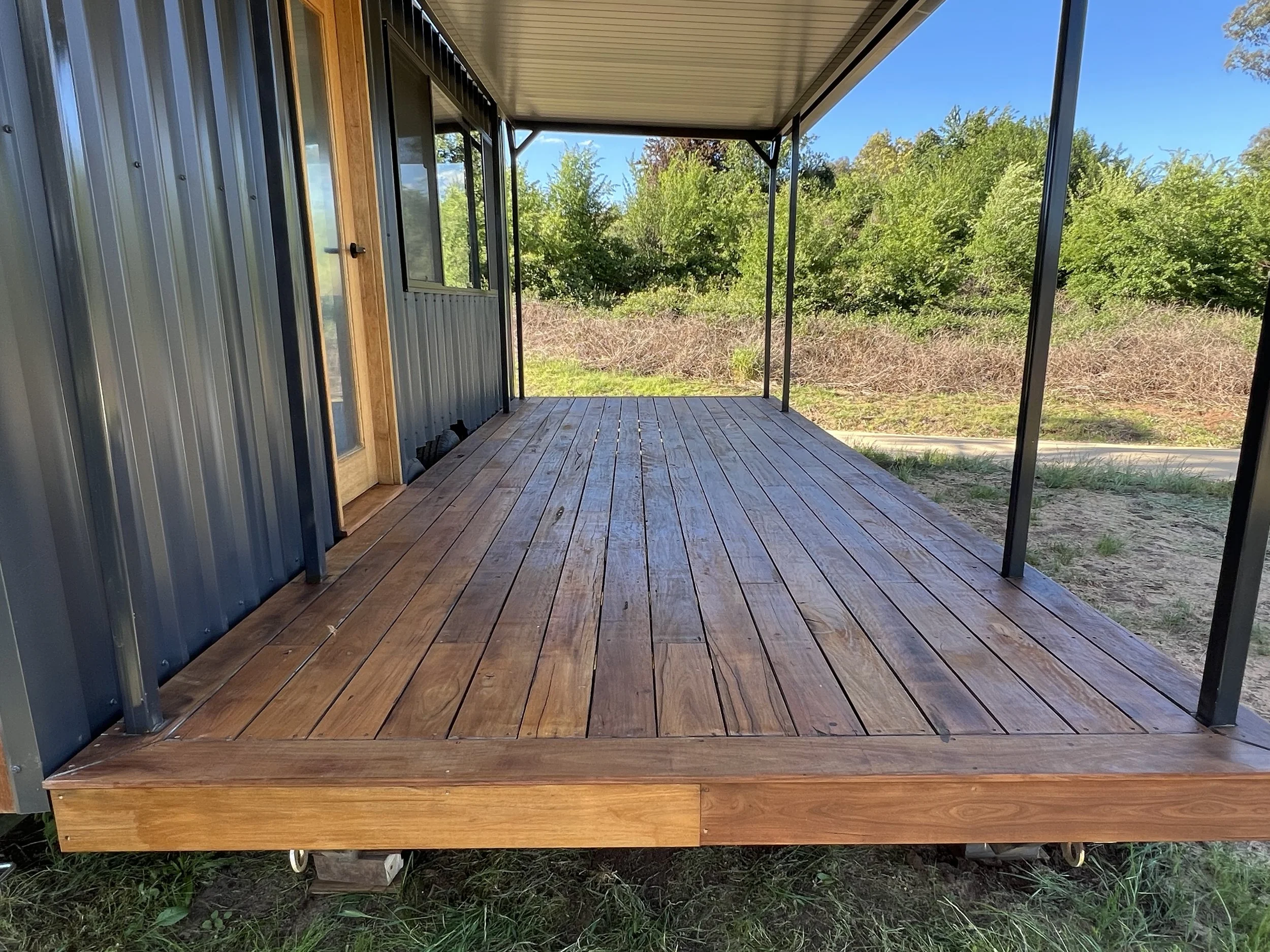 A newly built wooden porch with a metal roof and black metal supports, attached to a house with blue siding and a wooden door, surrounded by green trees and bushes.