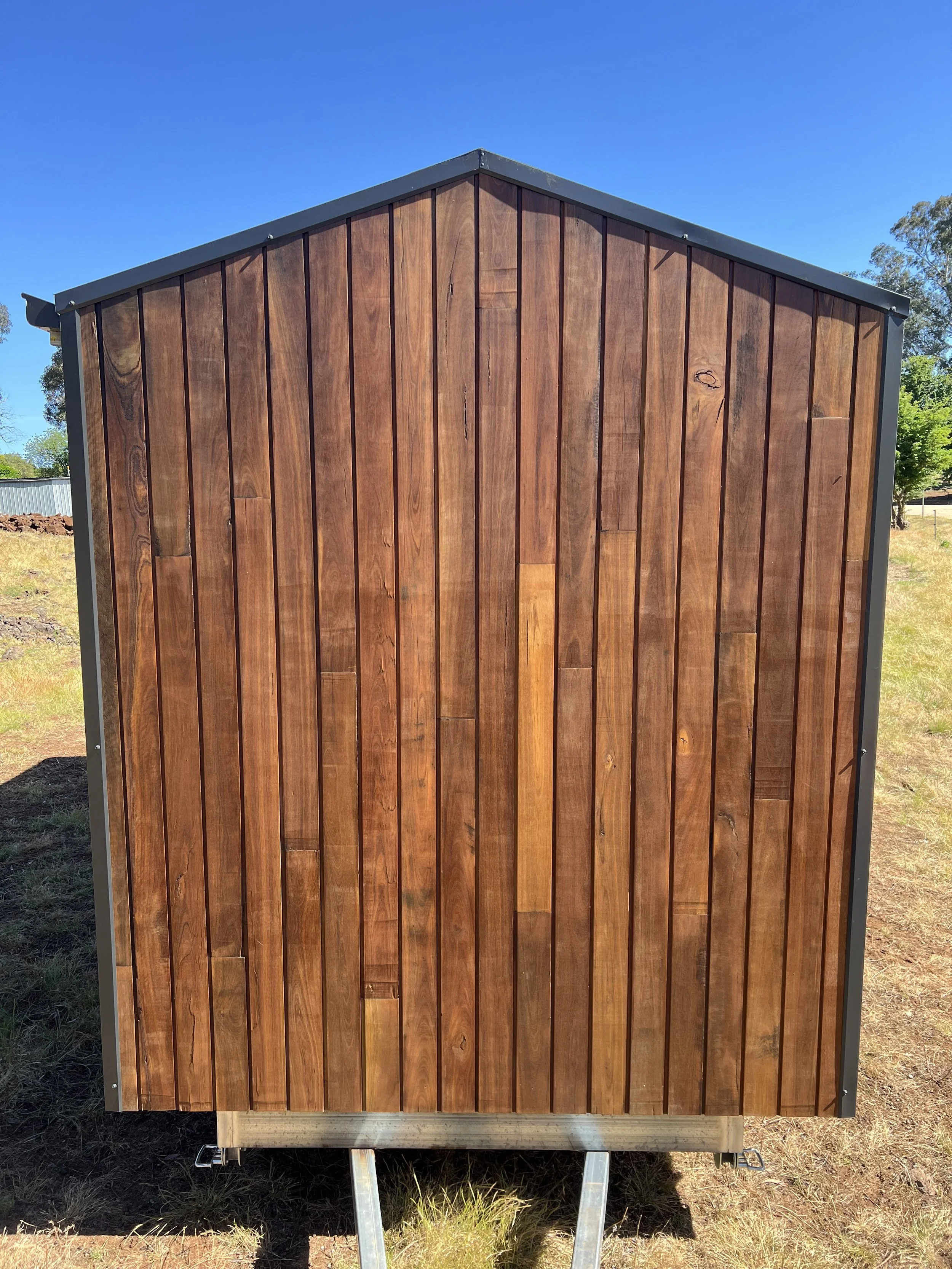Tiny house with a pitched roof, spotted gum shiplap cladding on a grassy lot under a clear blue sky.