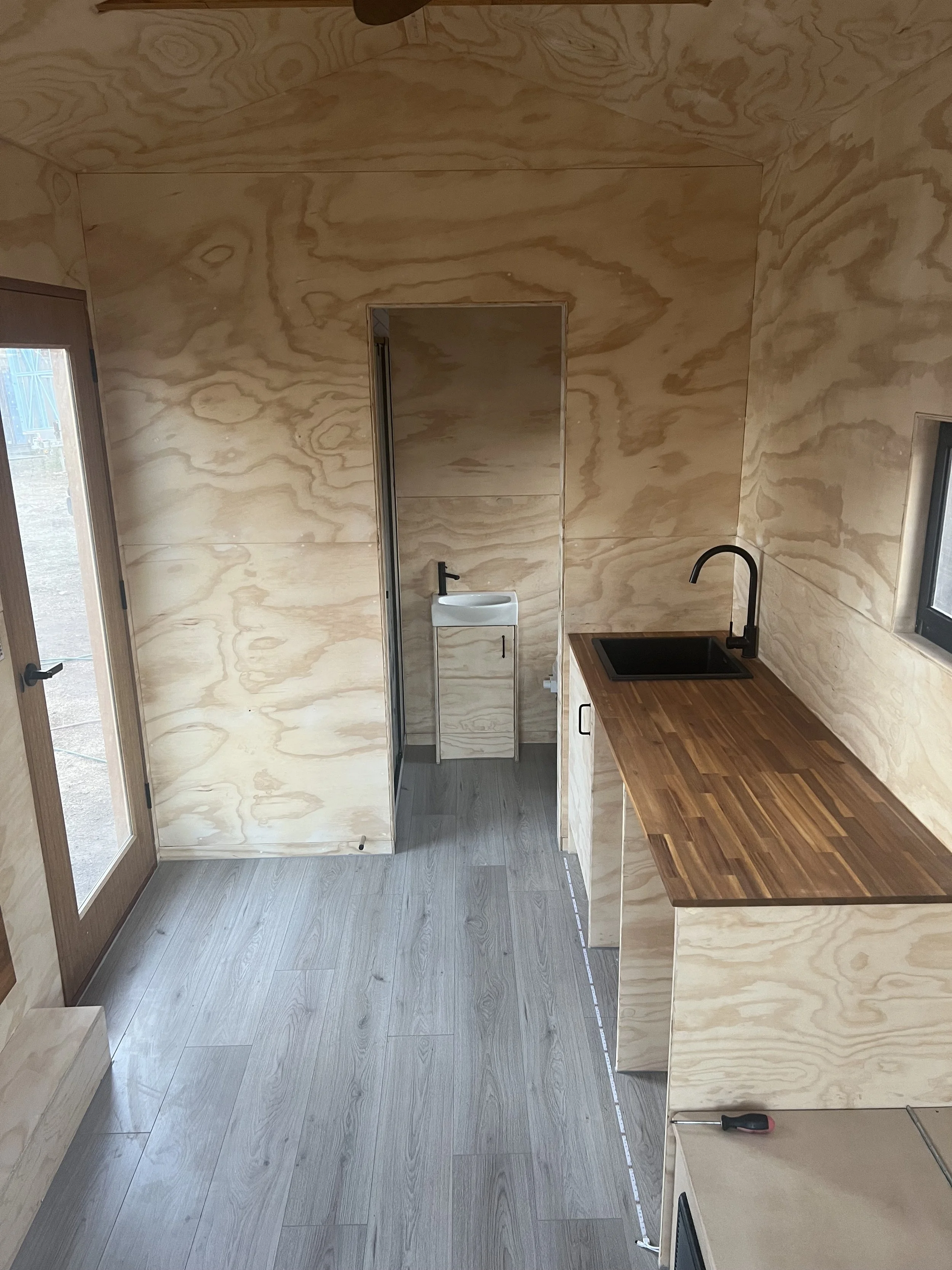 Interior of a small kitchen with plywood walls and a wooden countertop with a black sink, a small white laundry sink in the back, and a glass door on the left side.
