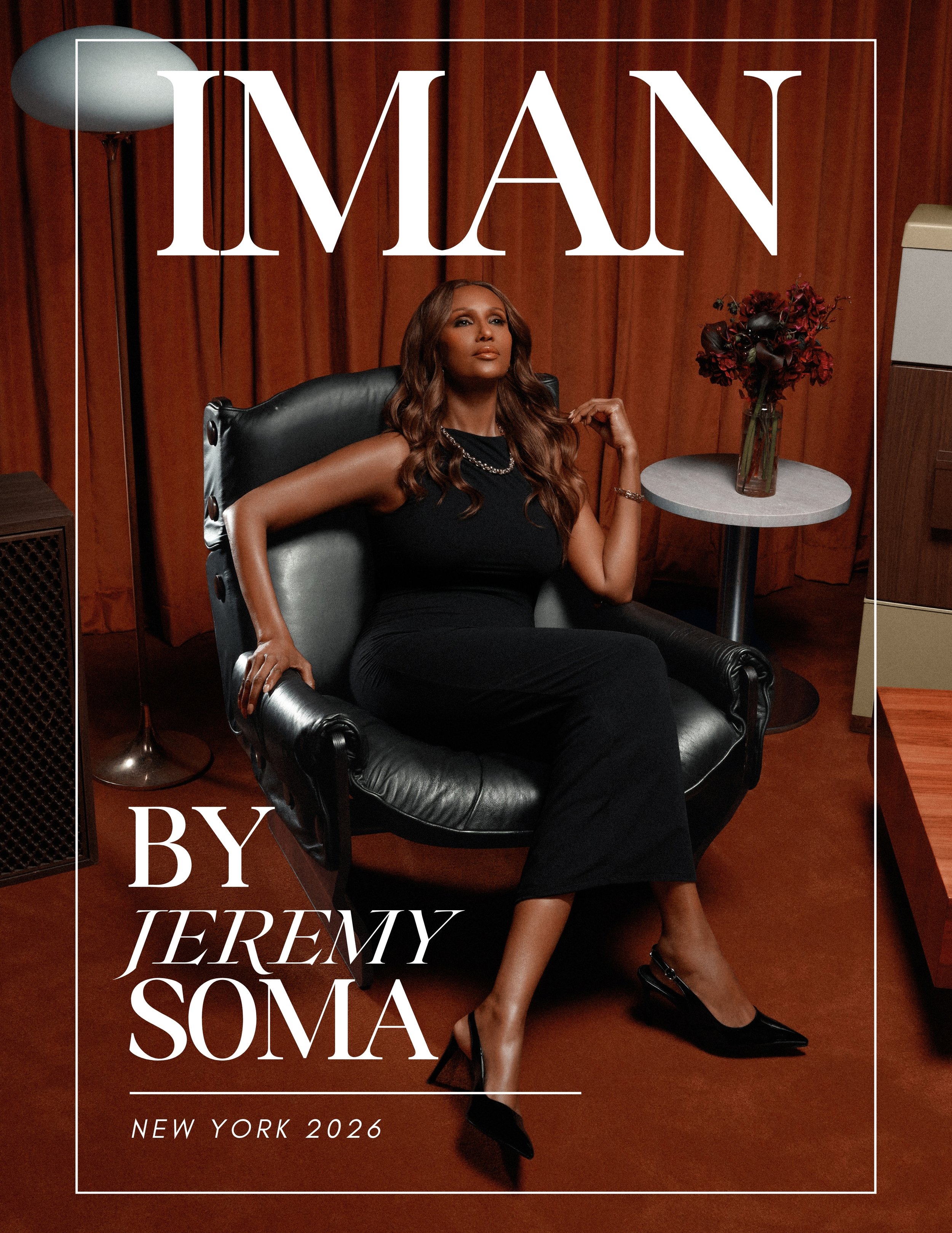 Cover of a magazine titled 'Iman' featuring super model Iman in a black dress seated on a black chair, with a flower vase on a side table, in a room with wooden paneling.