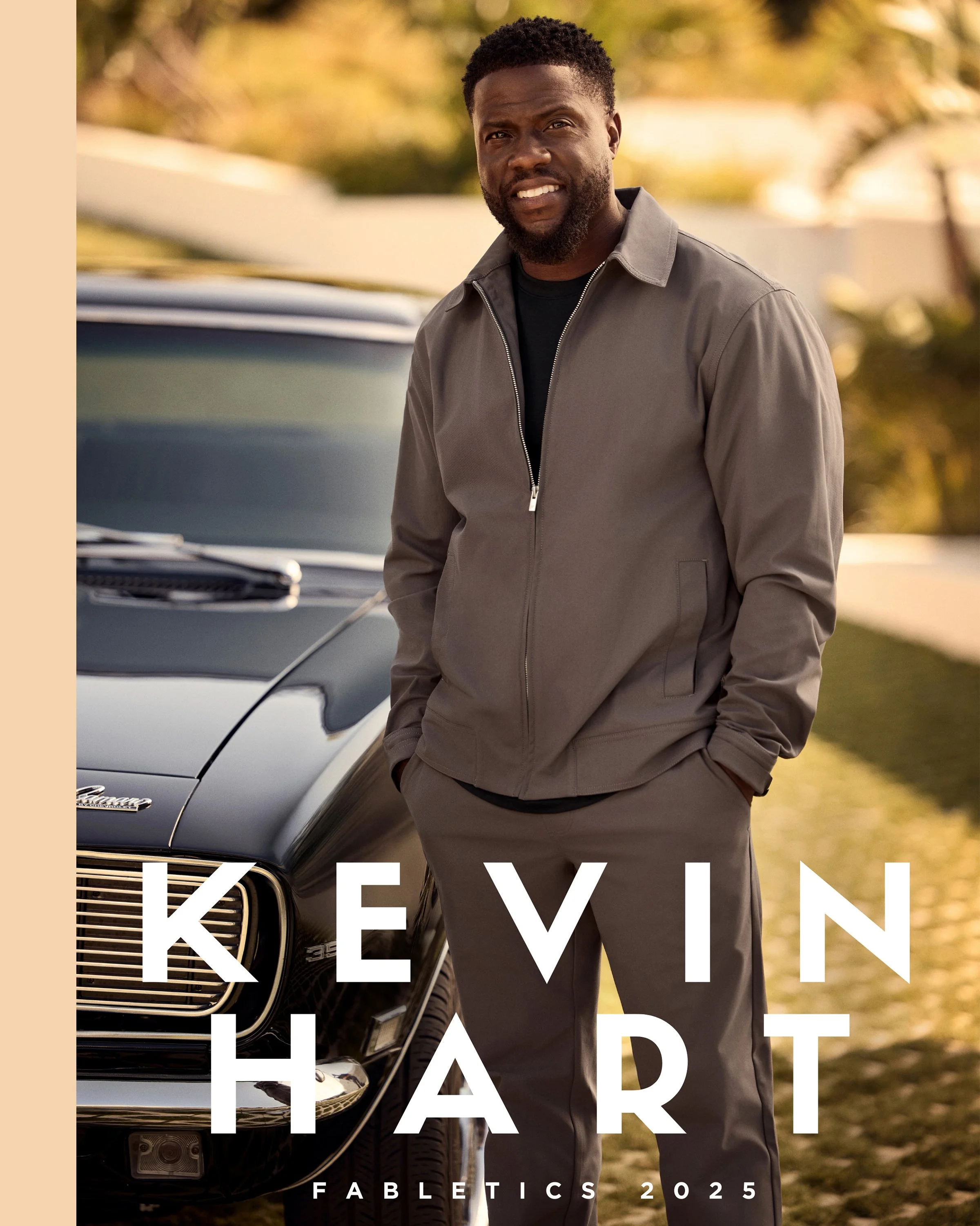 Cover photo of Kevin Hart for 'Fabletics 2025' featuring Kevin Hart standing outdoors next to a black vintage car, wearing a grey zip-up jacket and matching pants, with a blurred background of trees and sunlight.