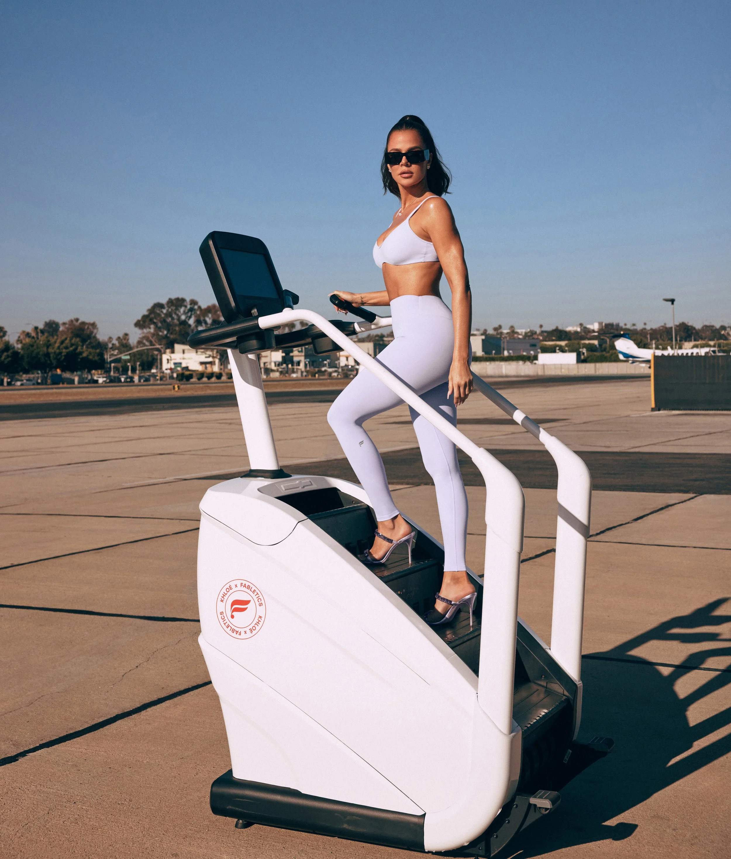 A woman dressed in white workout clothes and high heels standing on an airport treadmill machine outside, with sunglasses and holding a remote control.