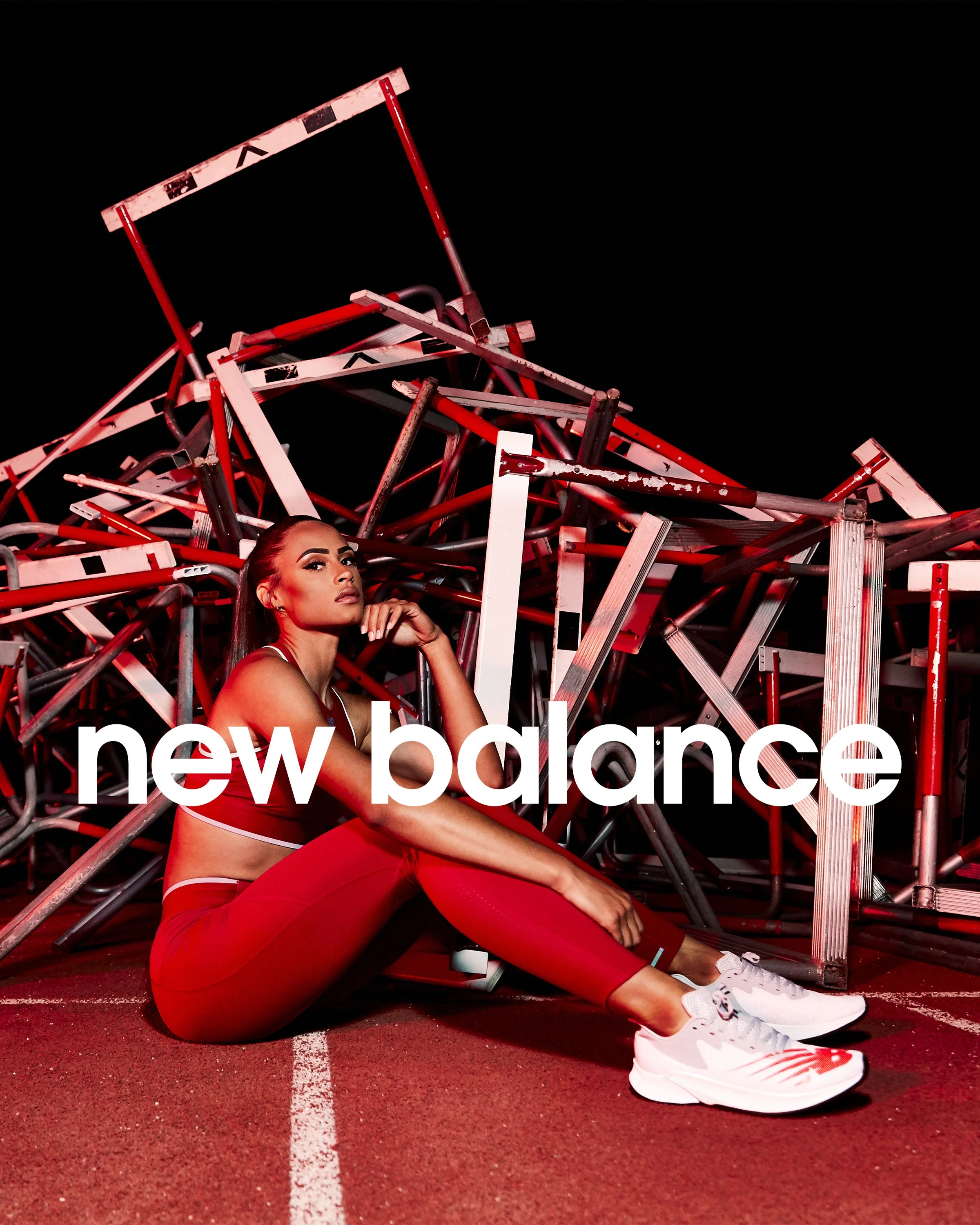 A woman in red athletic wear sitting on a track surface in front of a pile of metal hurdles, with the 'new balance' logo overlayed.