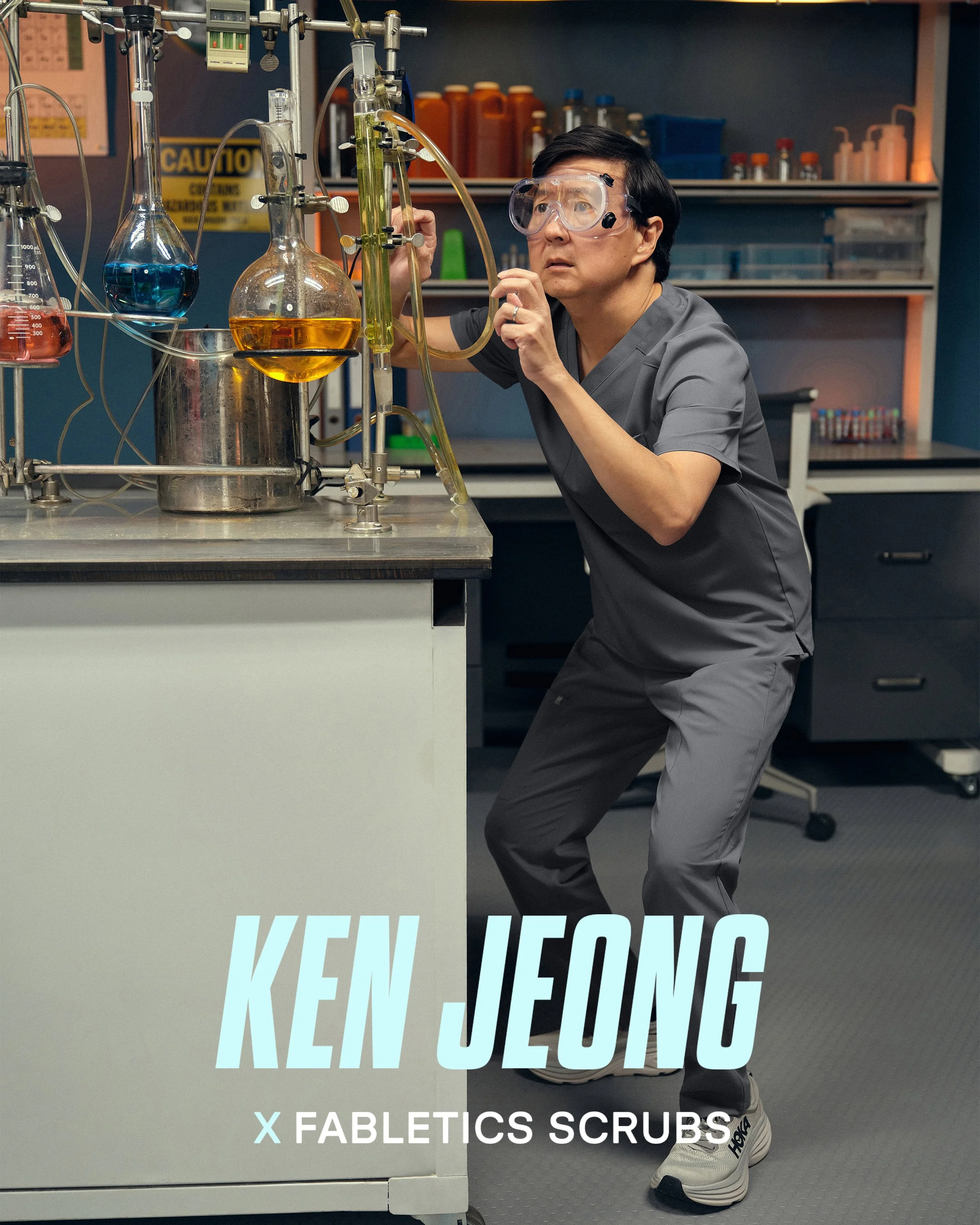 Ken Jeong in grey medical scrubs wearing safety goggles is leaning over a laboratory table with various glassware containing colorful liquids. He is in a science lab with shelves holding bottles and equipment in the background.
