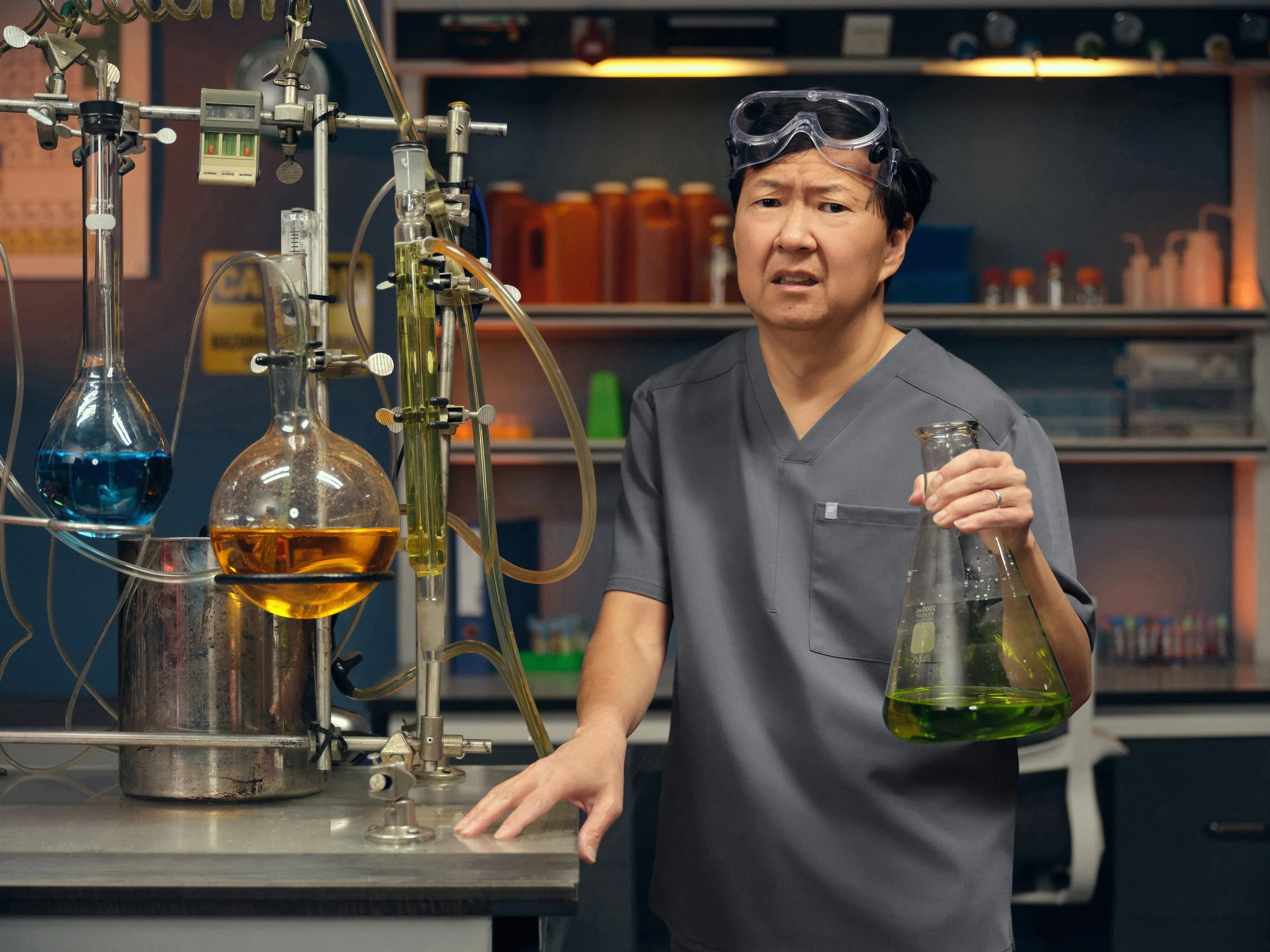 A man in a gray lab coat and safety goggles holding a glass beaker with a green liquid inside, standing in a chemistry laboratory with various glassware and equipment on the workbench.