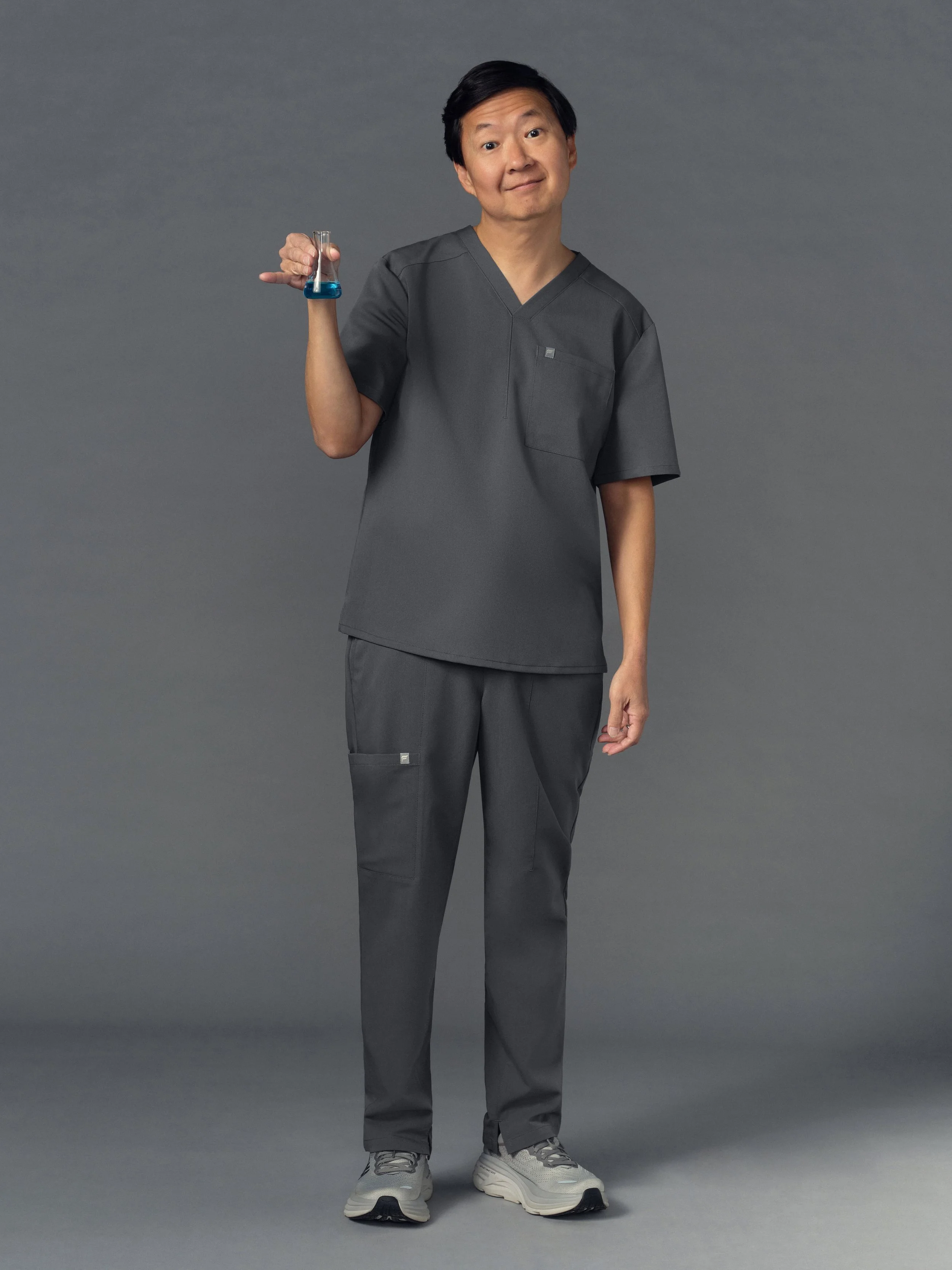 Person in gray medical scrubs holding a small test tube with blue liquid, smiling at camera against a gray background.