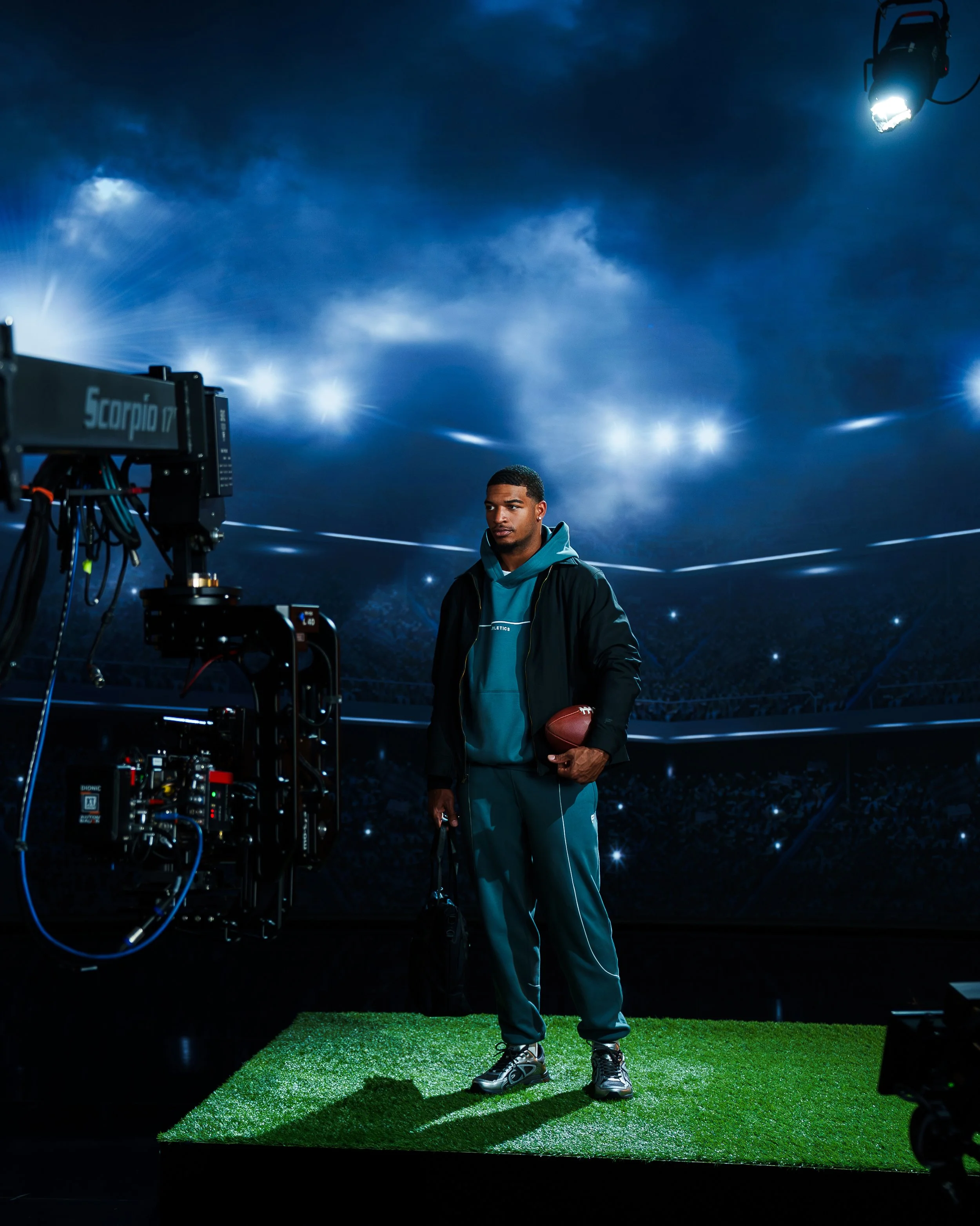 Young man in sportswear holding a football standing on a green platform in a stadium, filming equipment visible nearby, under dark skies and bright stadium lights.