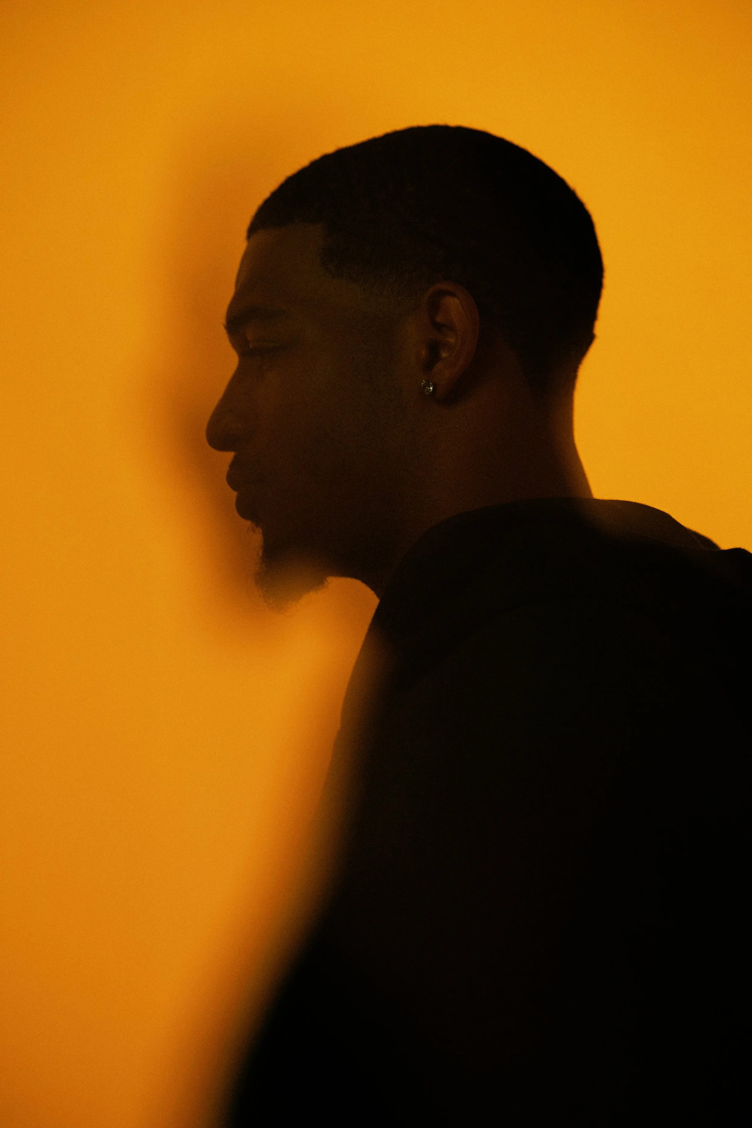 Silhouette of a young man with short hair and an earring, facing left against a warm orange background.