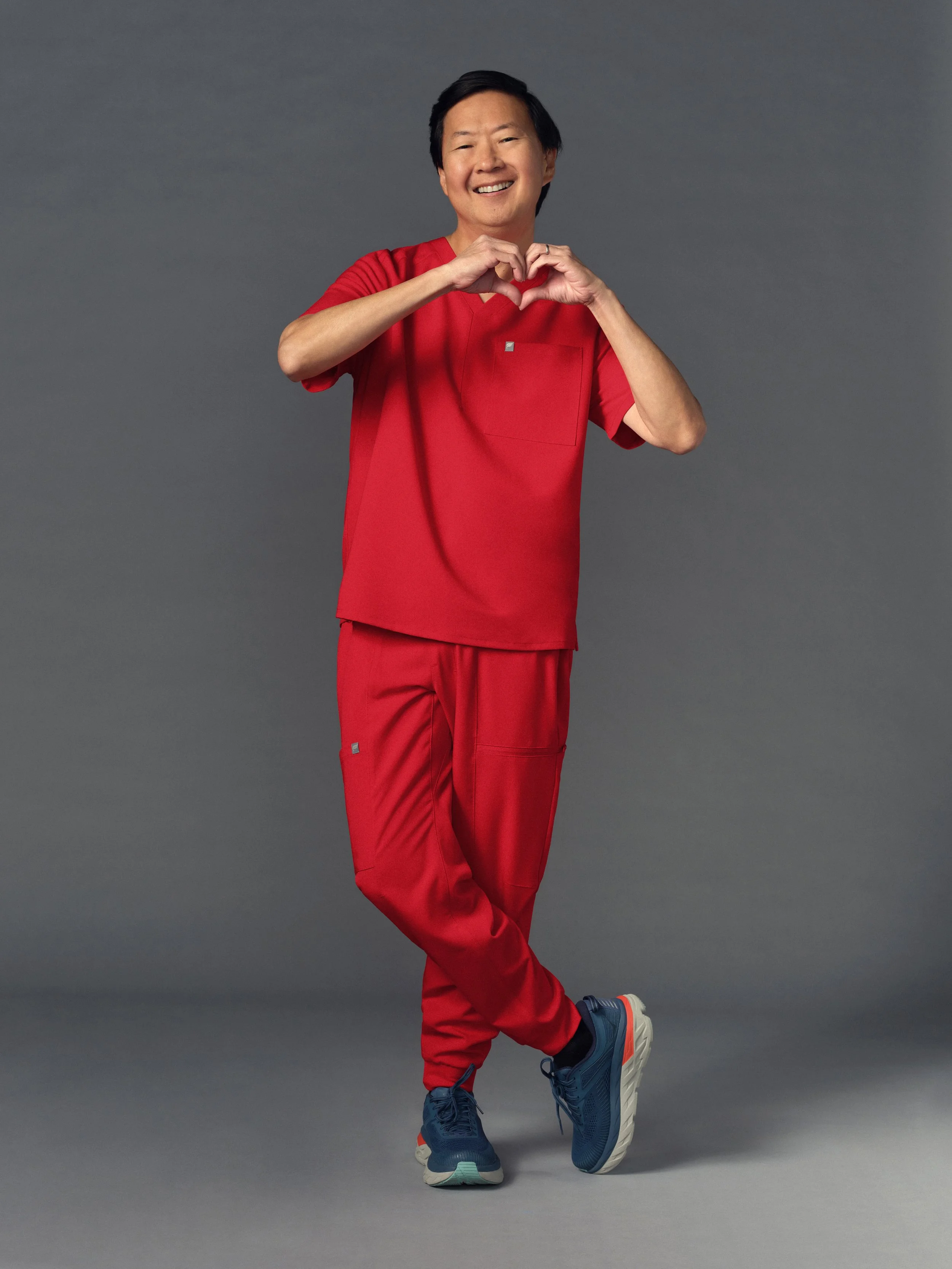 A man dressed in red scrubs standing against a gray background, making a heart shape with his hands and smiling.