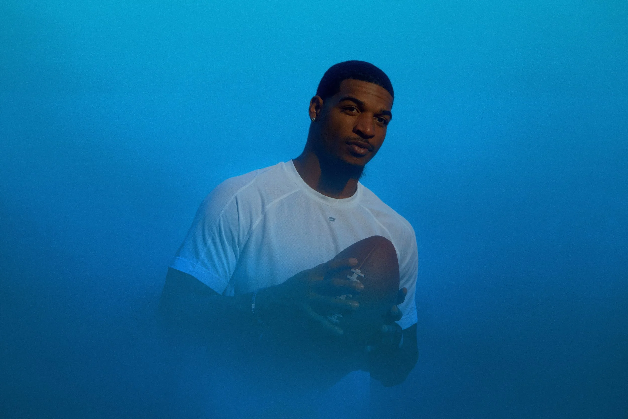 A young man with dark hair and earrings holding an American football, wearing a white athletic shirt, against a blue background.