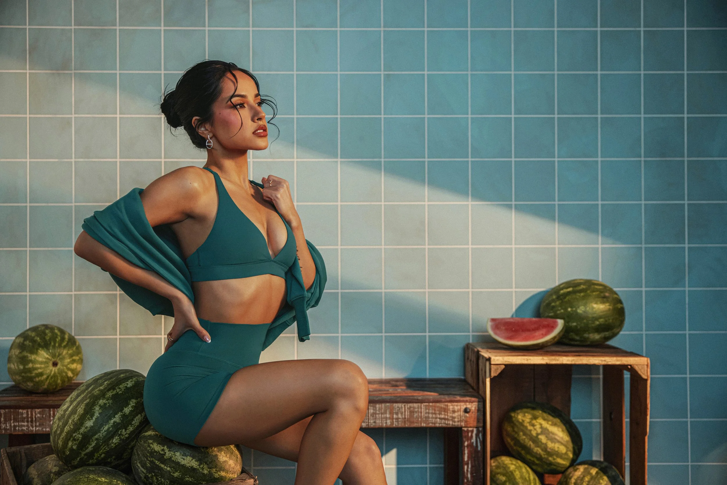 A woman in teal athletic wear poses against a tiled blue wall, surrounded by watermelons, with one cut open on a wooden stand.