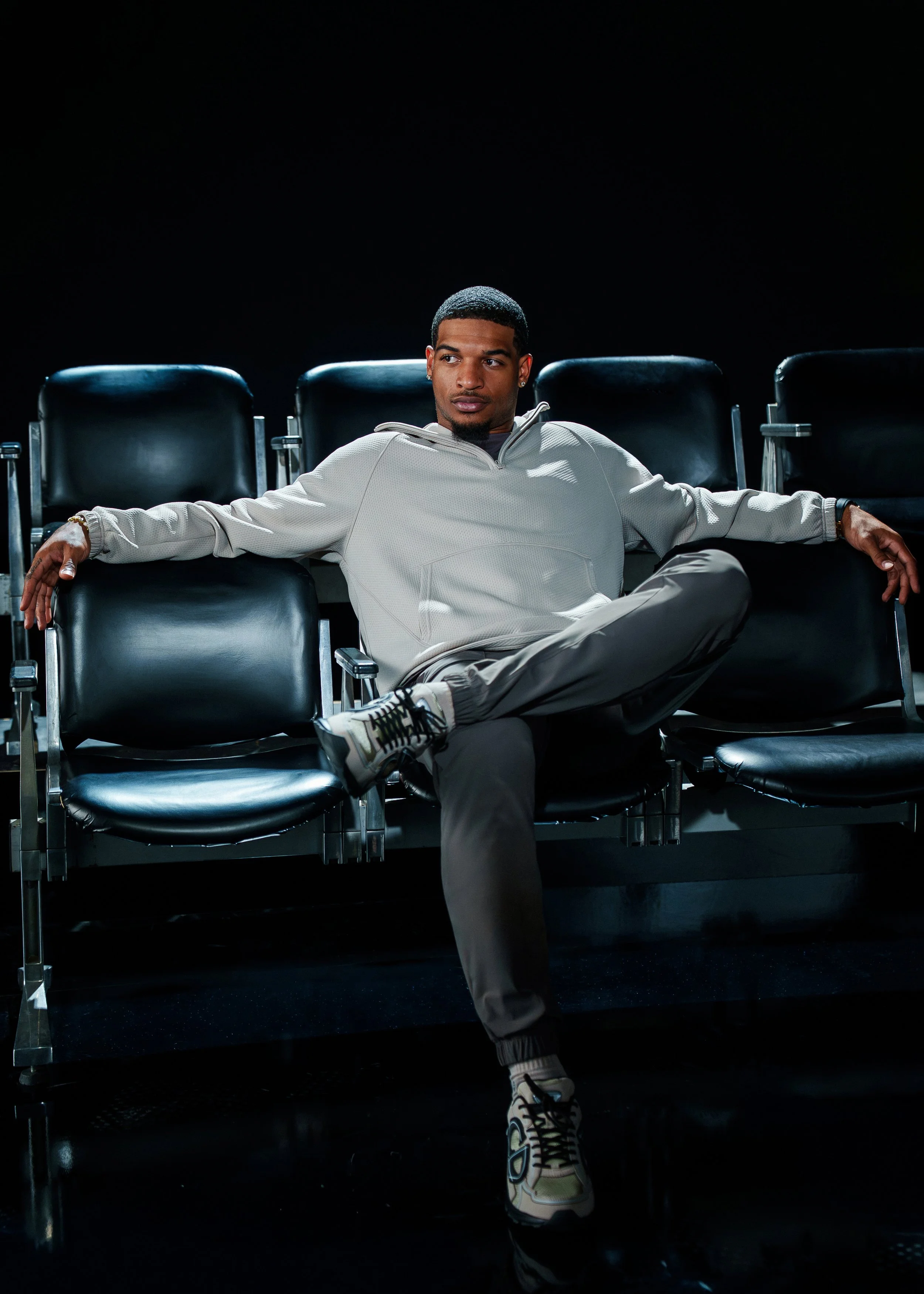 Young man sitting on a row of black chairs, wearing a gray tracksuit and sneakers, in a dimly lit room.