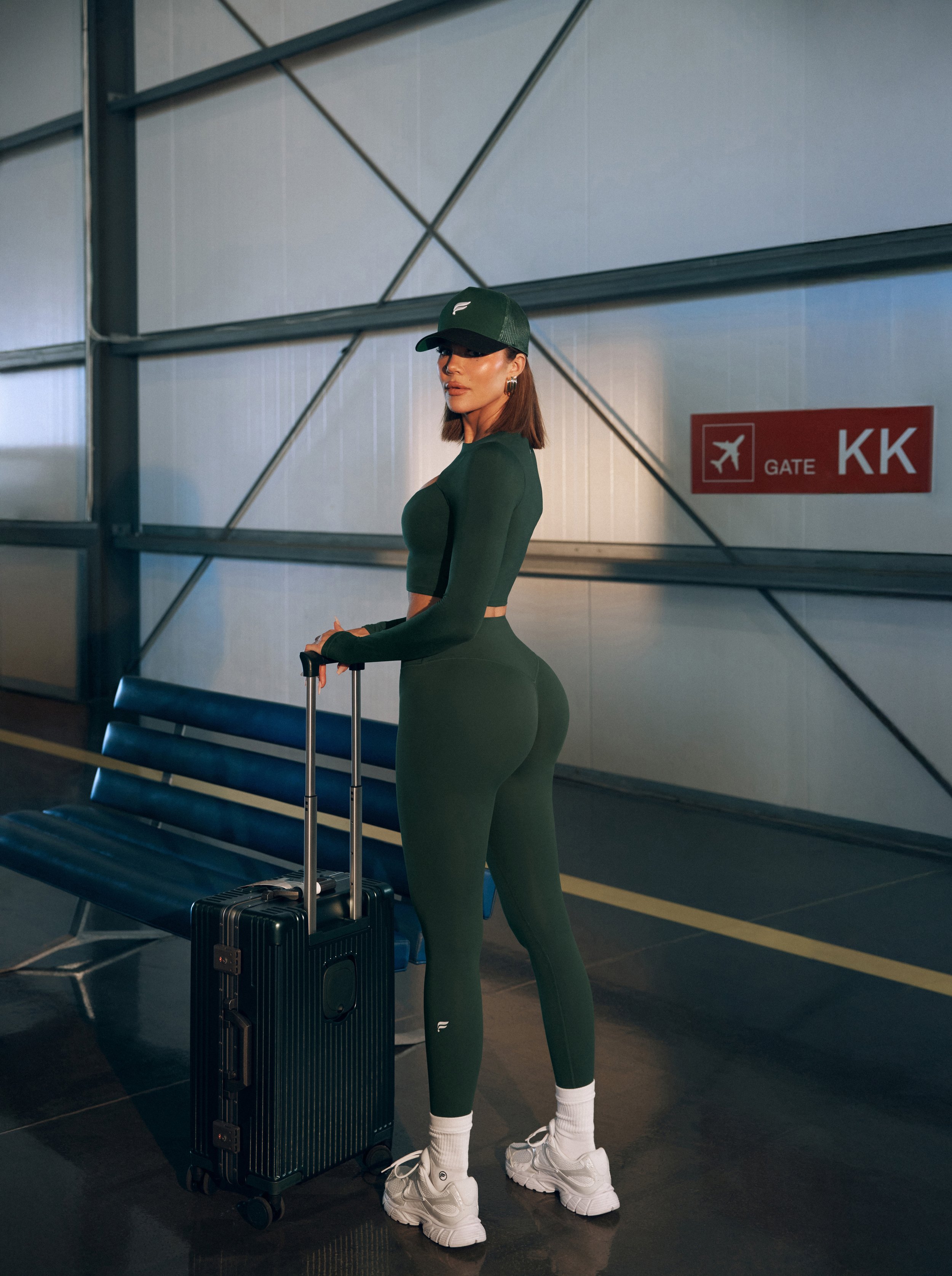 A woman in green athletic wear standing at an airport gate with a rolling suitcase, wearing a cap, white sneakers, and socks, looking sideways.