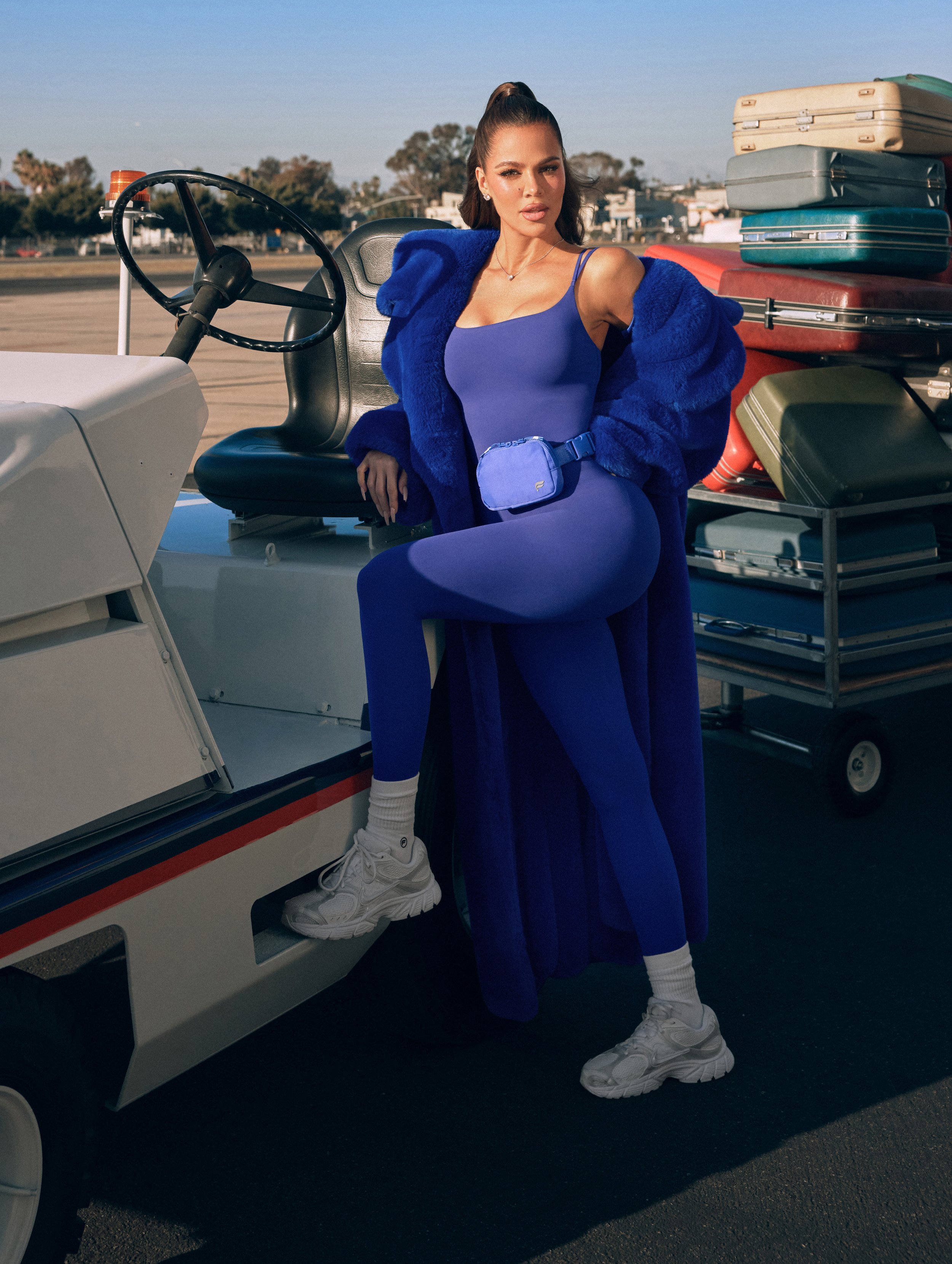 A woman in a blue outfit and white sneakers posing next to a small vehicle at an airport, with luggage stacked behind her.