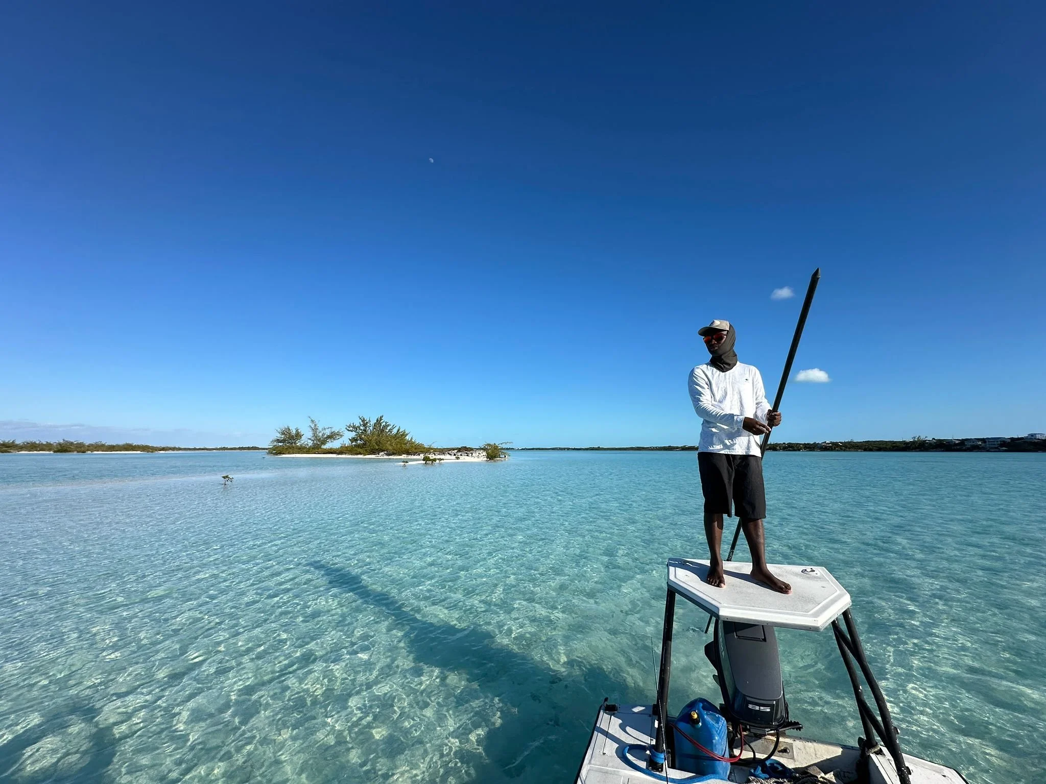 Person standing on a small boat holding a long pole in clear turquoise water with a small island and blue sky in the background.