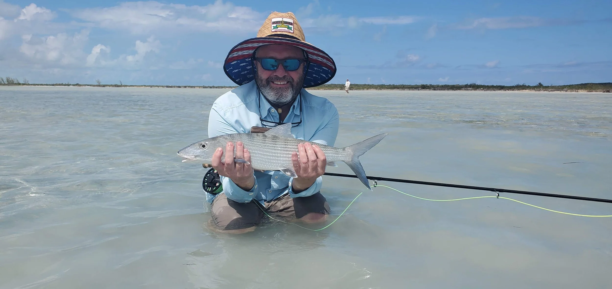 A man kneeling in shallow water holds a fish with both hands, wearing a large sun hat, sunglasses, a light blue button-up shirt, and khaki shorts. He is smiling and appears to be fishing in a coastal or estuarine environment.