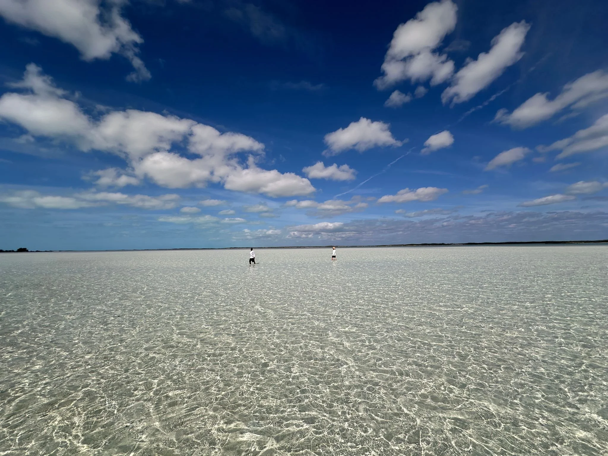 A vast flat, white salt flat under a partly cloudy blue sky with two people walking in the distance.