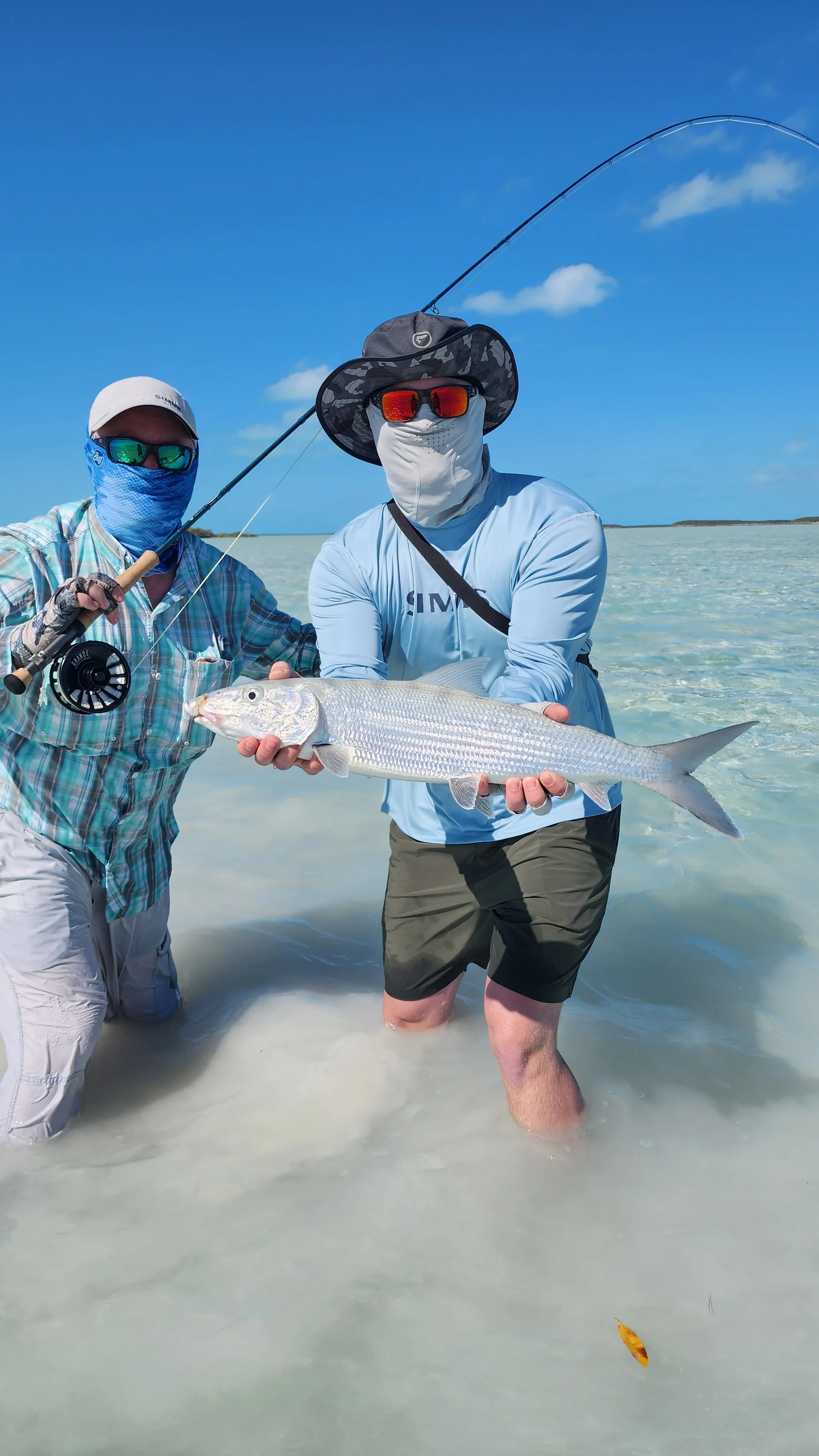 Two men in fishing gear holding a large fish on a sandy beach with clear blue sky.