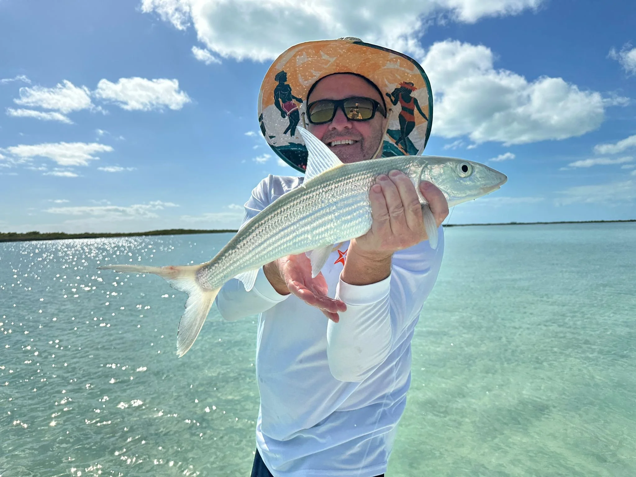 Man with sunglasses and a wide-brimmed hat holding a fish in a shallow, clear body of water on a sunny day with blue sky and clouds.