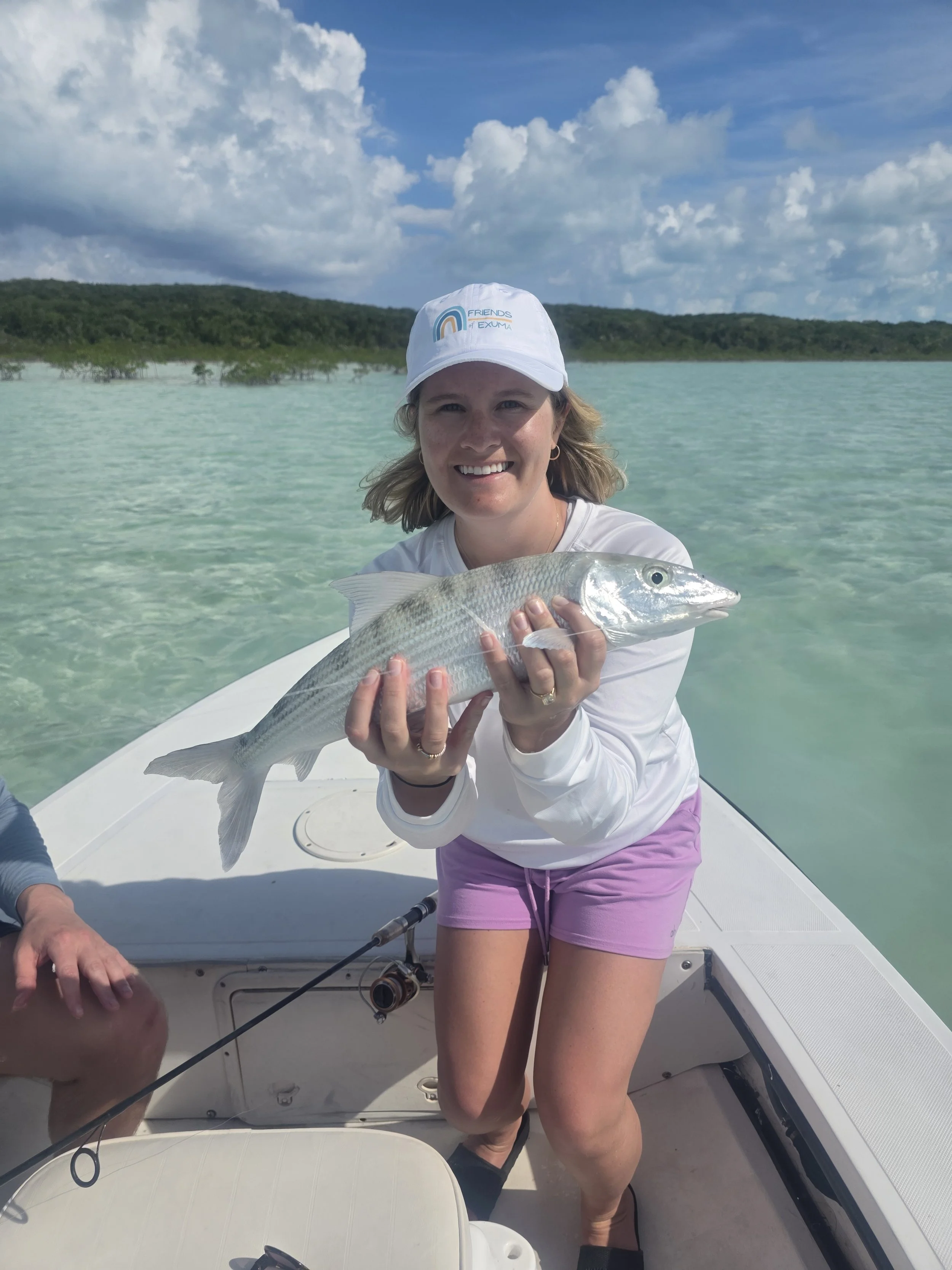 Woman in white long-sleeve shirt, purple shorts, and white hat holding a large striped fish on a boat in clear turquoise water, with green hills and sky with clouds in the background.