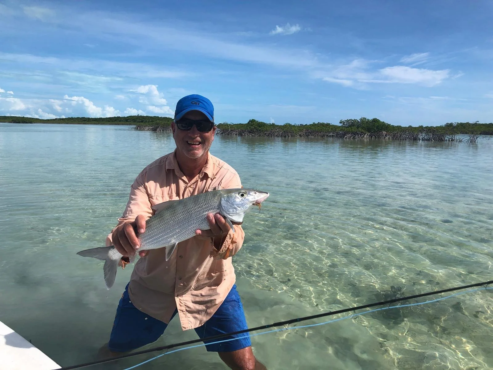 Smiling man wearing sunglasses, blue cap, and beige shirt holding a large fish, standing knee-deep in clear water with a beach and green trees in the background.