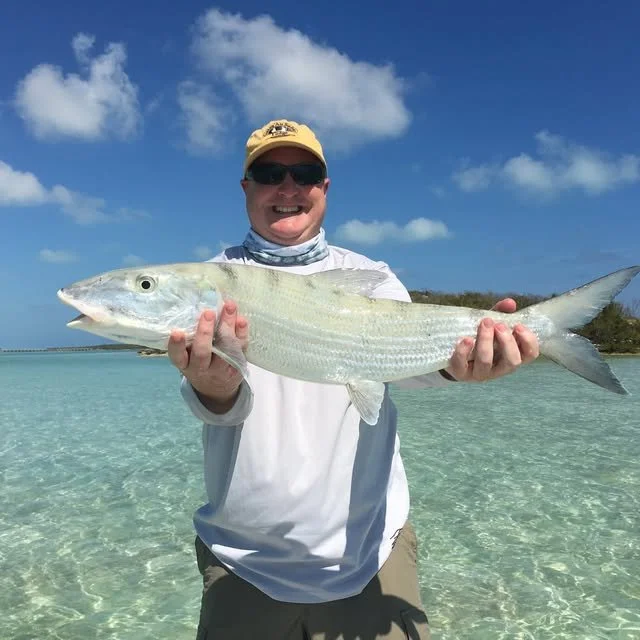 A man wearing sunglasses, a beige cap, and a white shirt holding a large fish on a clear, shallow beach with a blue sky and some clouds in the background.
