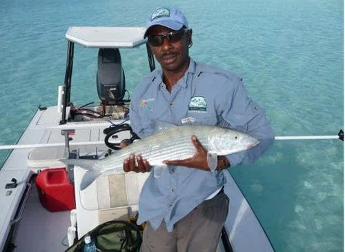 Man in fishing attire holding a large fish on a boat in clear blue water.