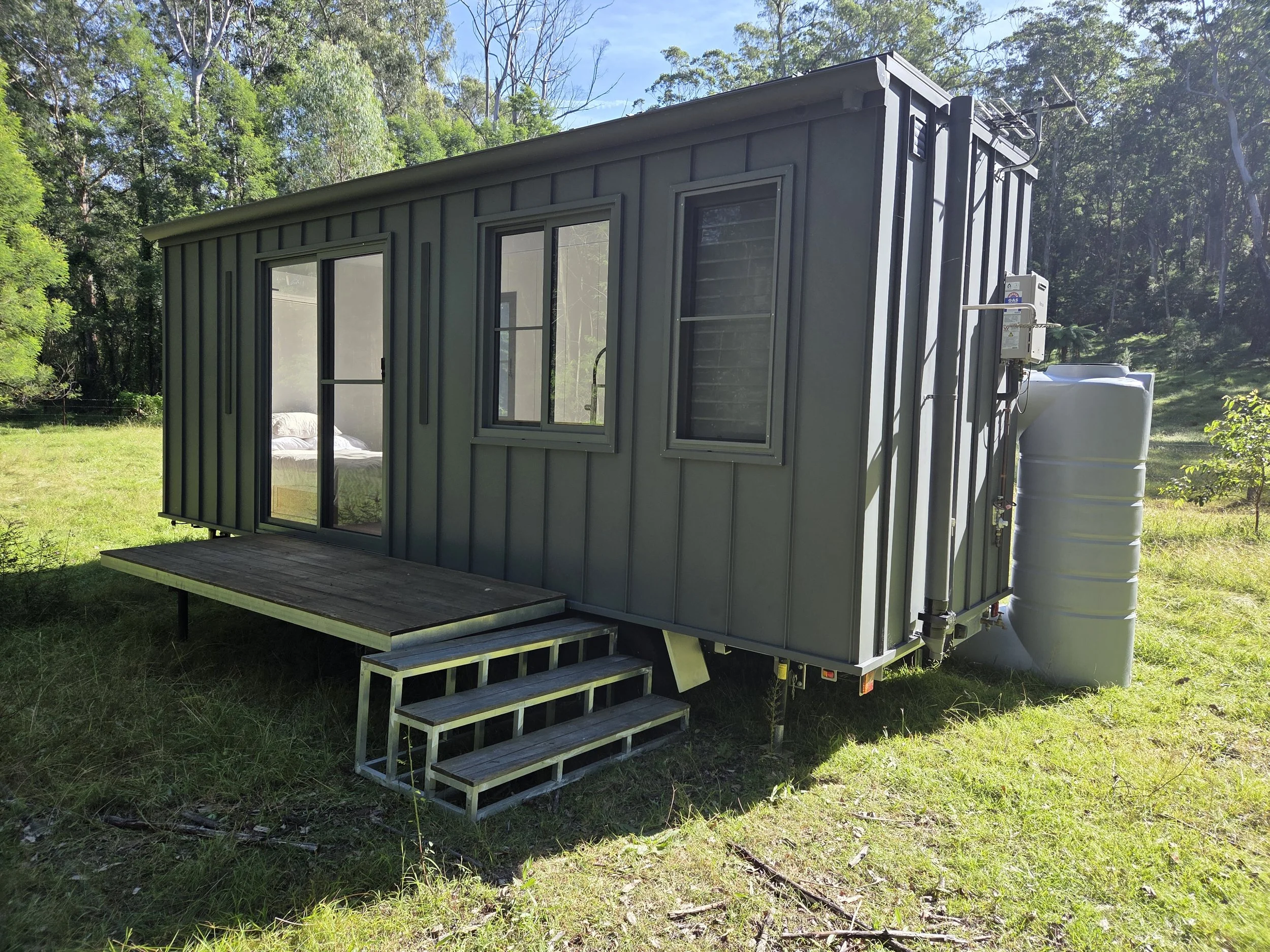 A small gray tiny house with a sliding glass door and two windows, built on a raised wooden platform with steps, situated on a grassy area surrounded by trees under a clear sky.