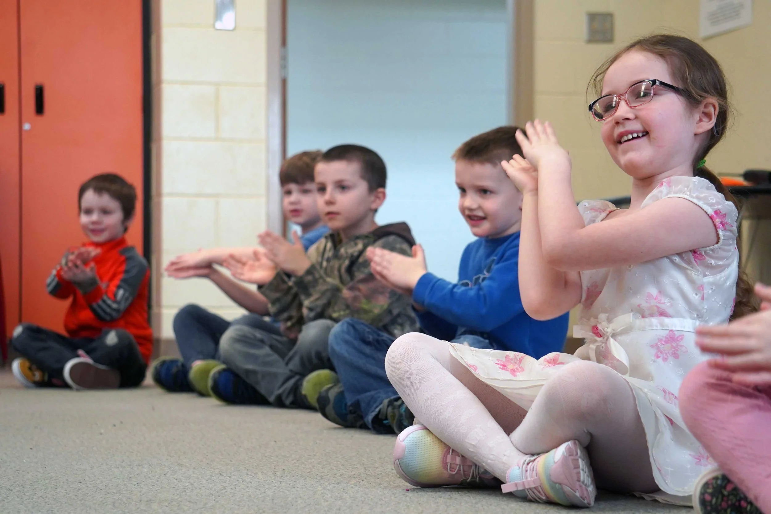 Group of young children sitting on the floor in a classroom, clapping and smiling.
