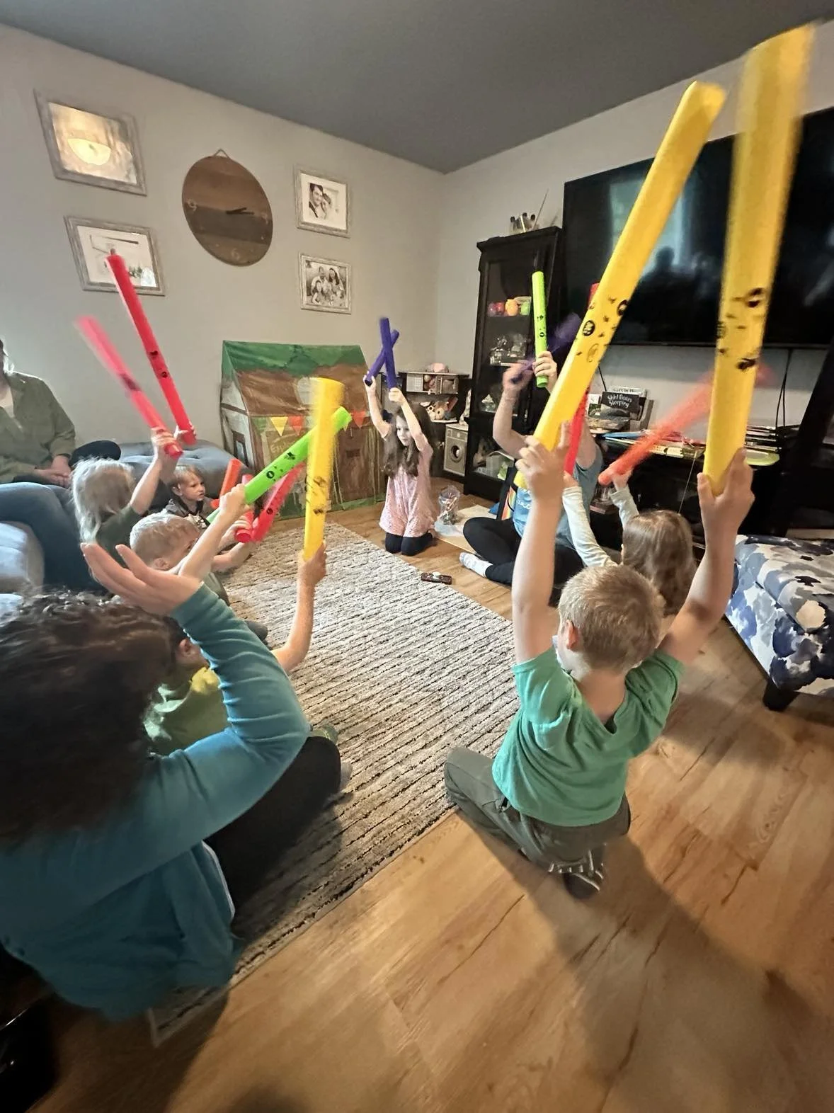 Children sitting on the floor with adult, holding colorful pool noodles, during a birthday party in a living room.