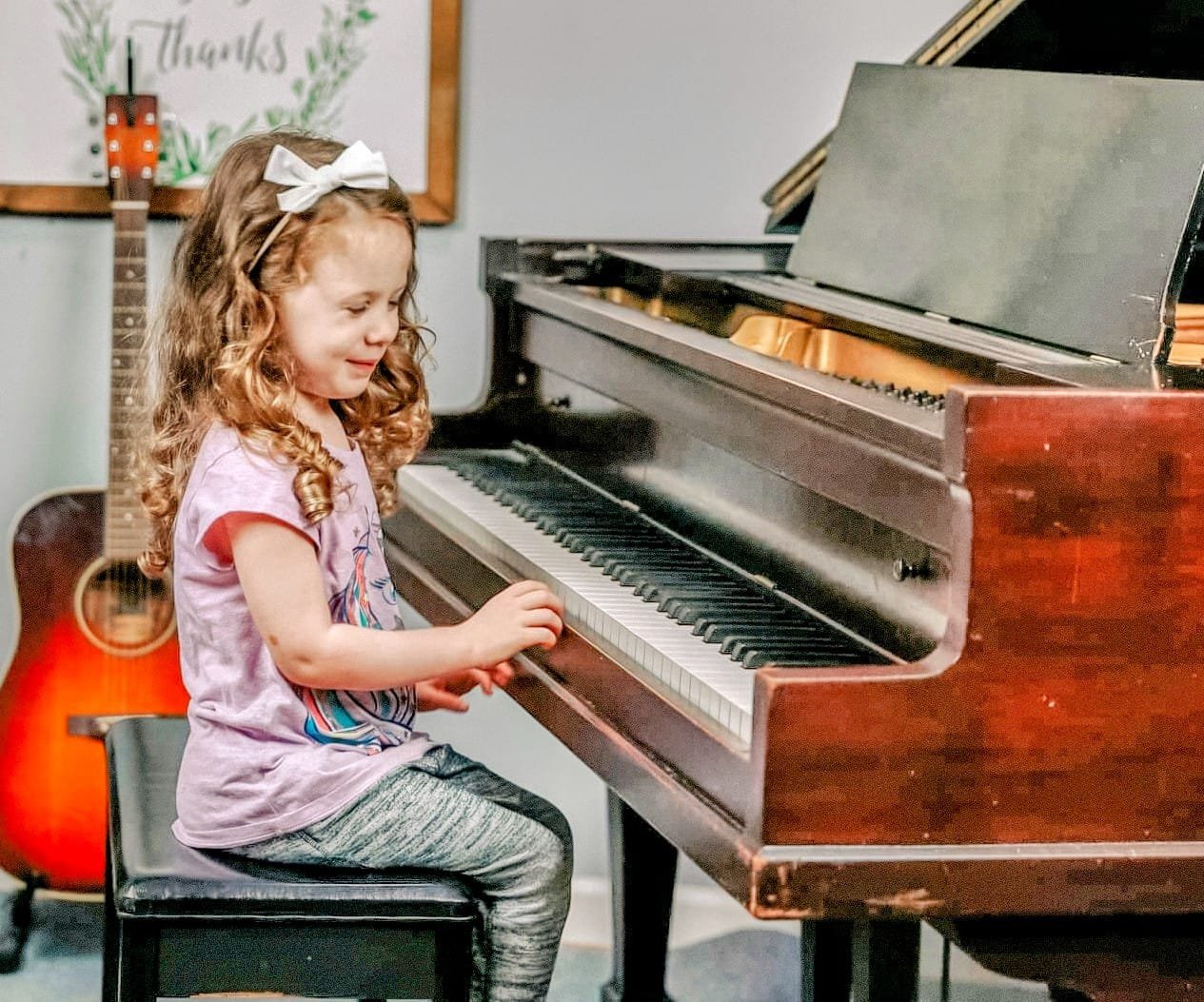 A young girl with curly red hair, wearing a pink shirt and gray leggings, playing a brown upright piano, with a guitar standing behind her and a thankful sign on the wall in the background.