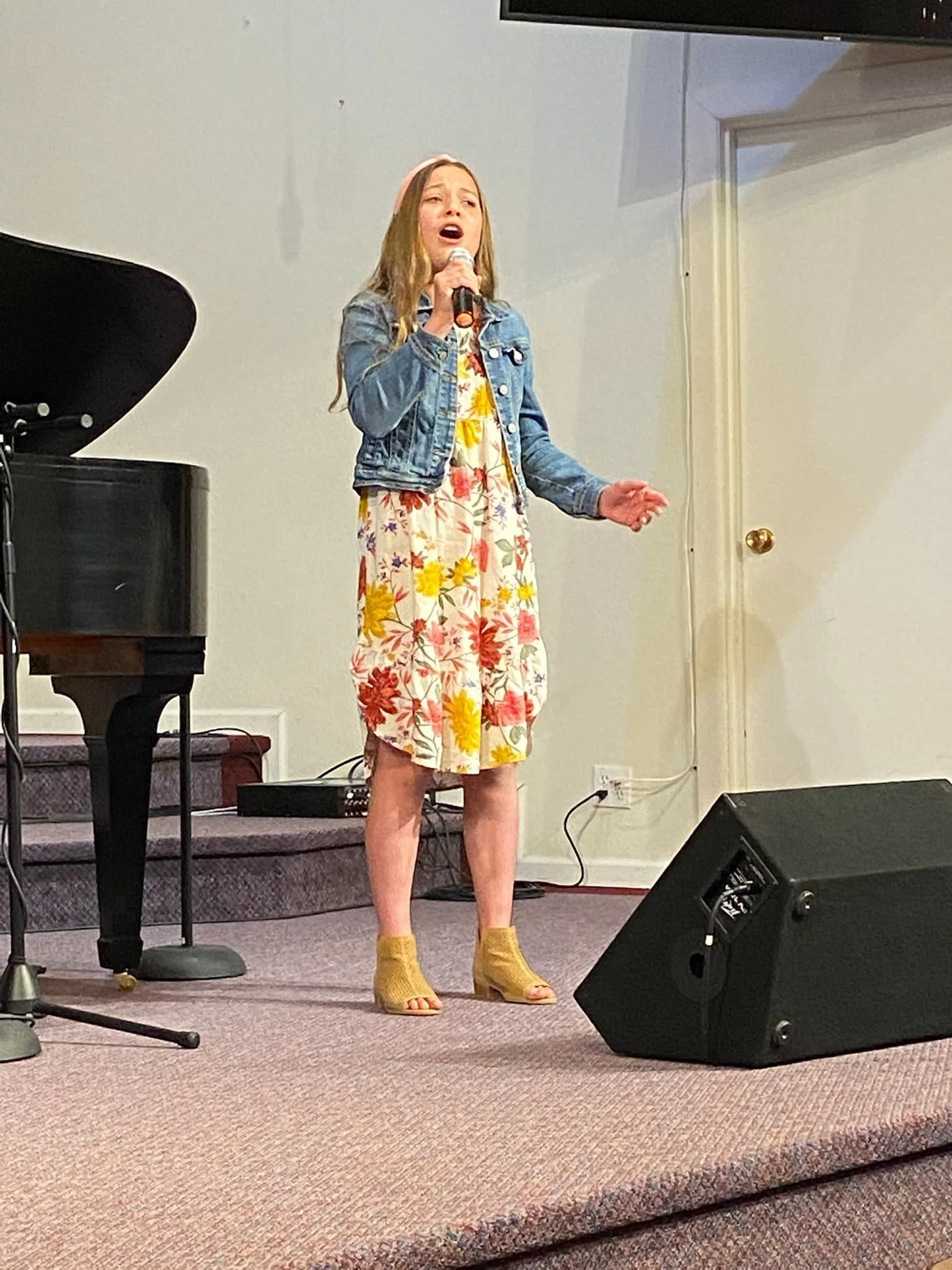 A girl singing into a microphone on a stage with a grand piano nearby. She is wearing a floral dress, yellow open-toe shoes, and a denim jacket.