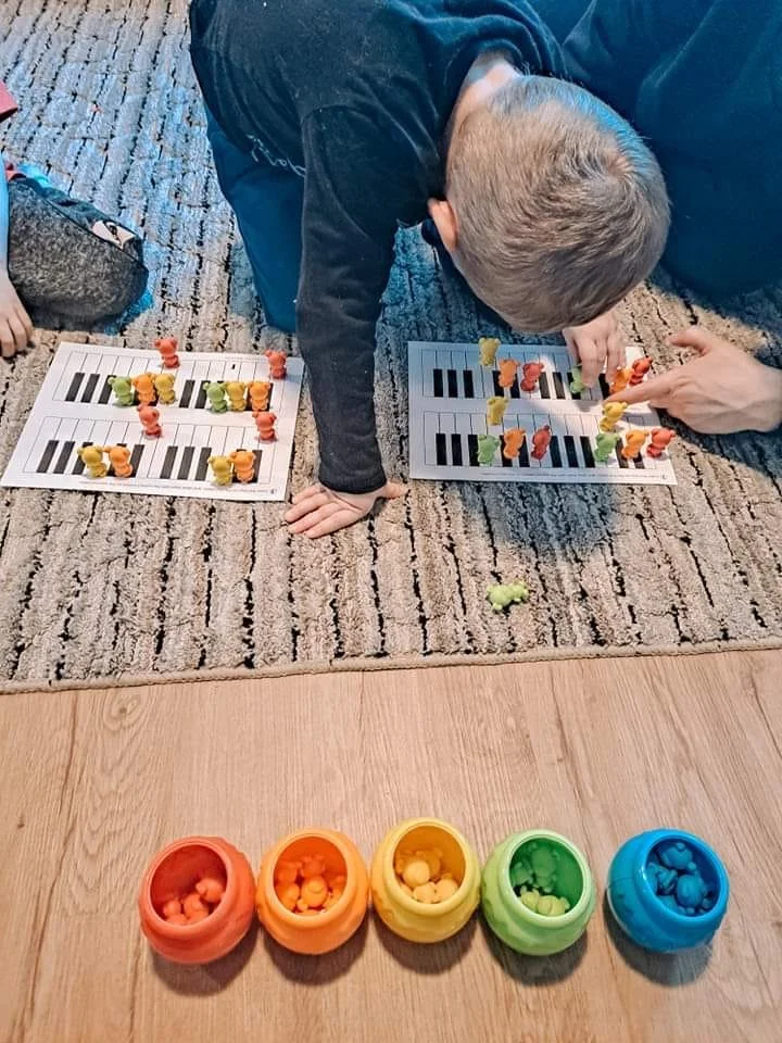 A young child playing a game with small animal-shaped game pieces on paper game boards on a carpeted floor, with containers of additional game pieces in various colors in front of him.