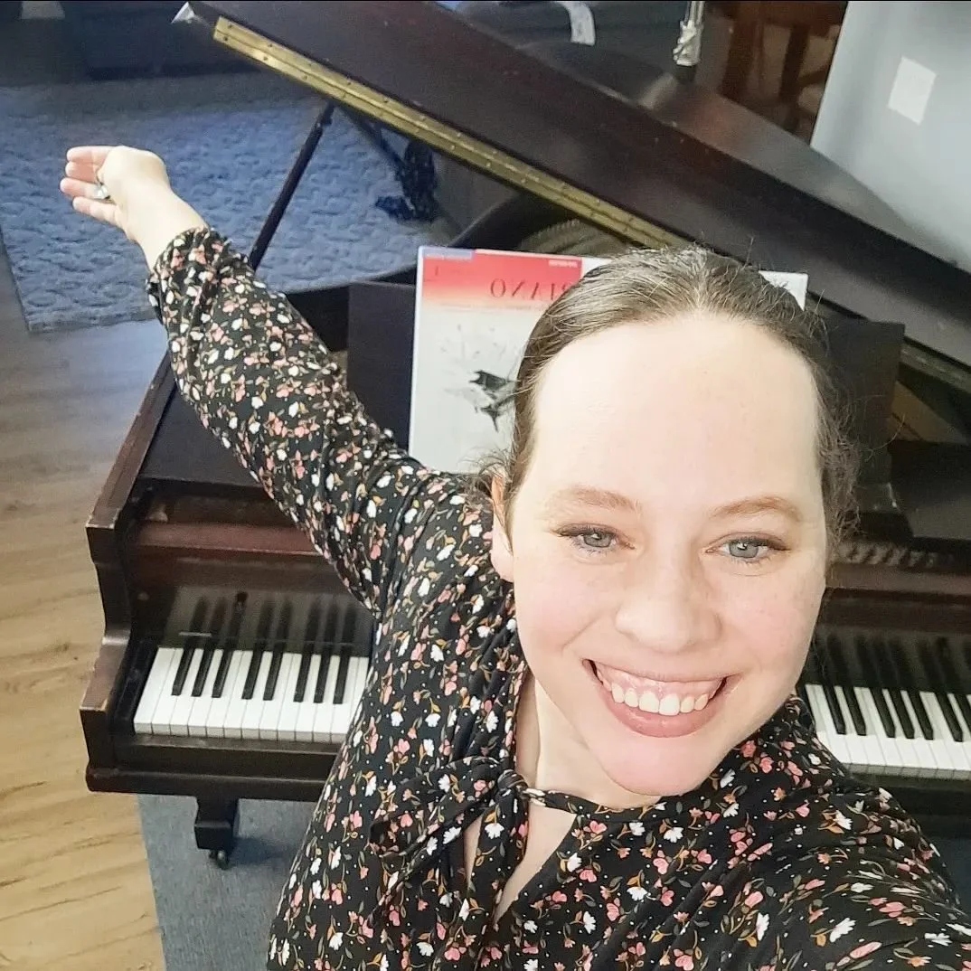 A woman smiling in front of a grand piano, taking a selfie with her arm extended. She is wearing a black floral dress, and there is a music book on the piano.