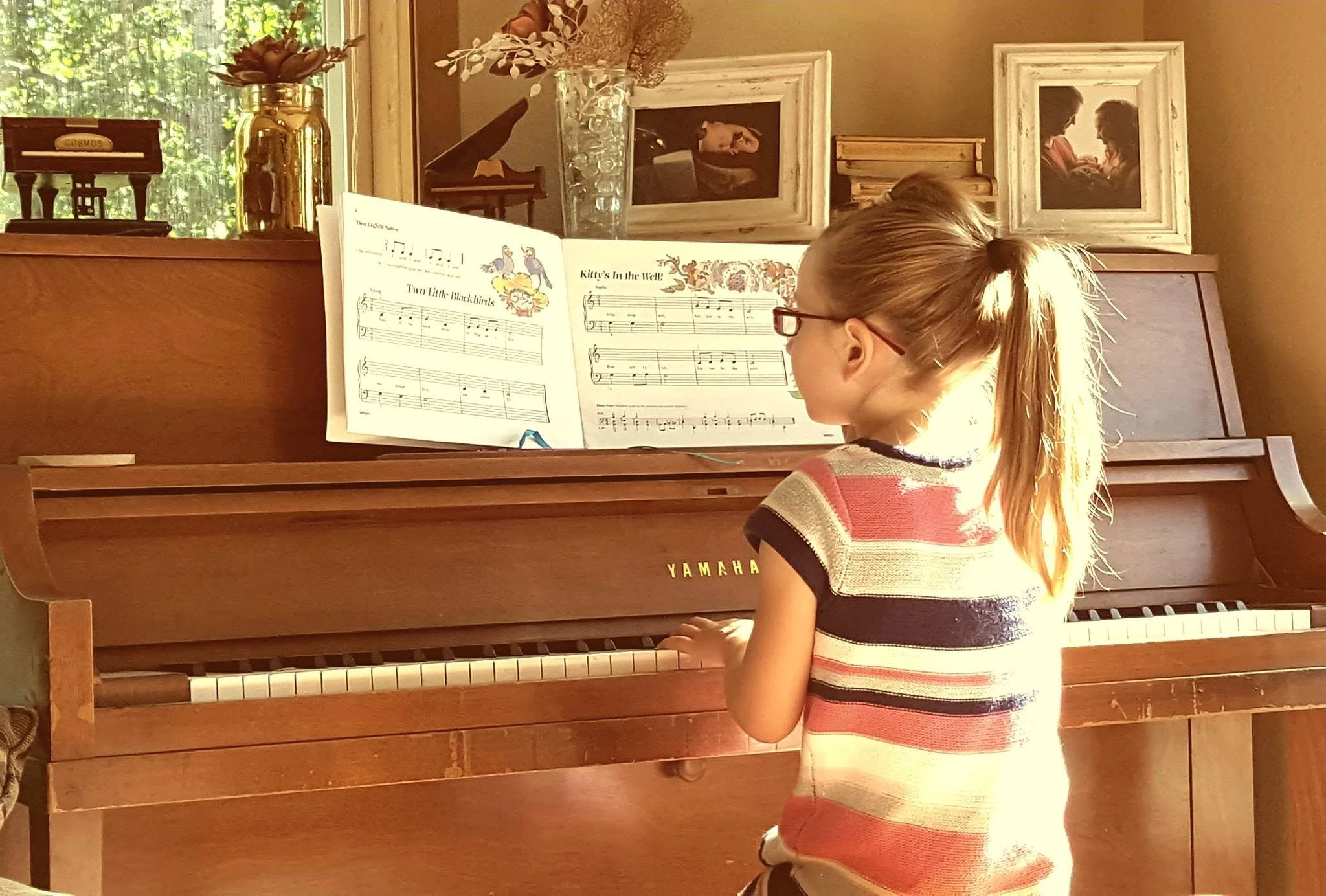 Young girl with glasses practicing piano in a cozy room with framed pictures on the wall and a window with sunlight.