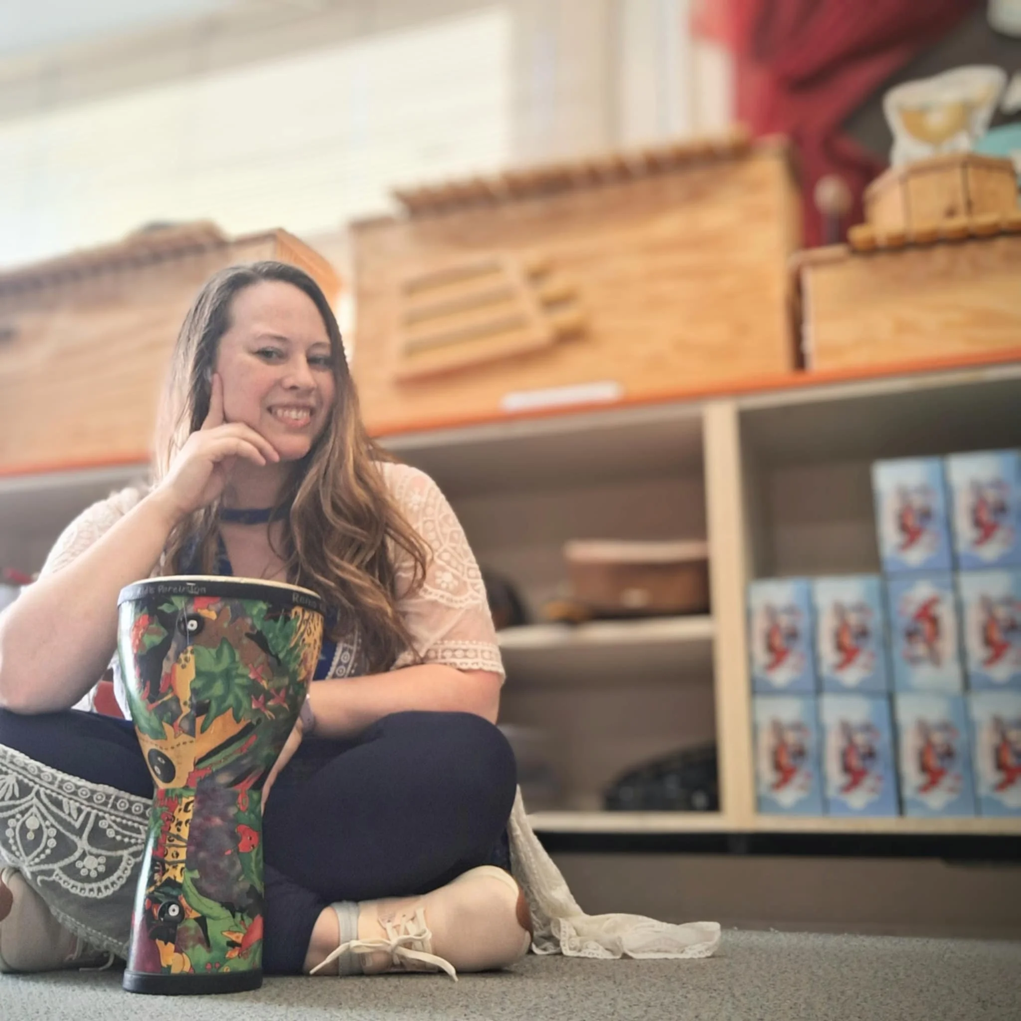 A woman with long hair smiling, sitting on the floor with her legs crossed, in front of a colorful decorative ceramic vase, inside a store with wooden shelves and boxes in the background.