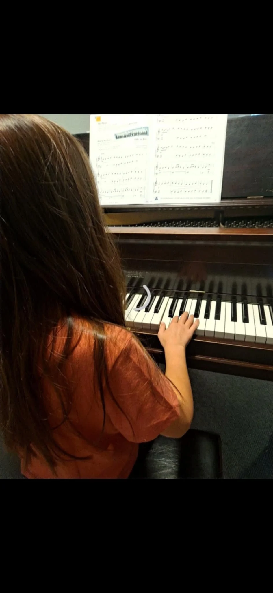 A young girl with long brown hair playing a brown upright piano, reading sheet music placed on the music stand.