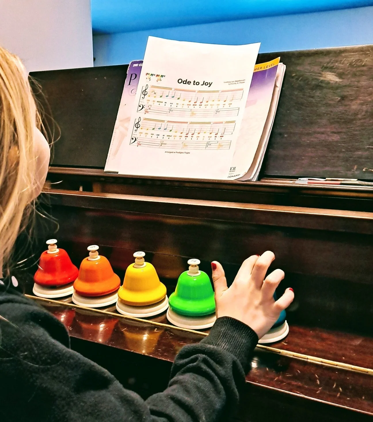 A person playing a colorful xylophone with black, red, orange, yellow, and green bells while reading music sheet titled "Ode to Joy" on a piano.