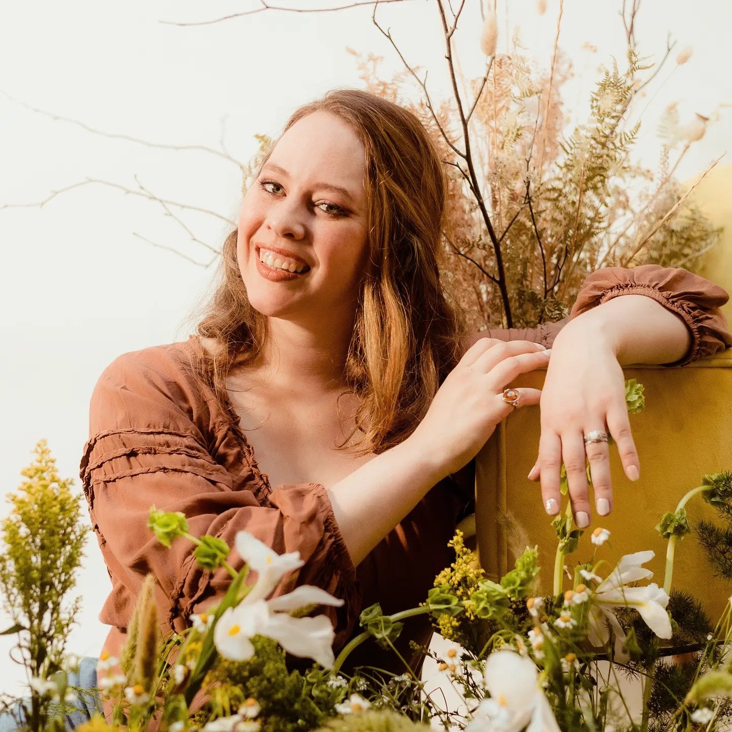 A smiling woman with light brown hair and wearing a brown blouse, leaning on a yellow container with her hand showing rings, surrounded by flowers and plants outdoors with a background of trees and an overcast sky.