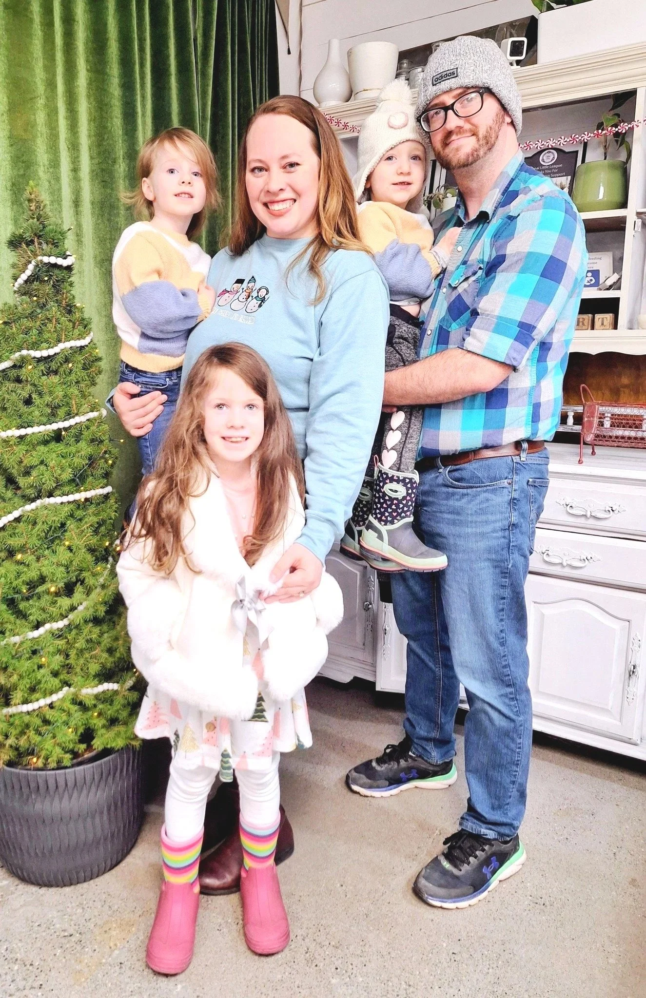 A family of six posing indoors near a decorated Christmas tree, with a green curtain in the background. The family includes a woman, a man, and four children, all smiling at the camera.