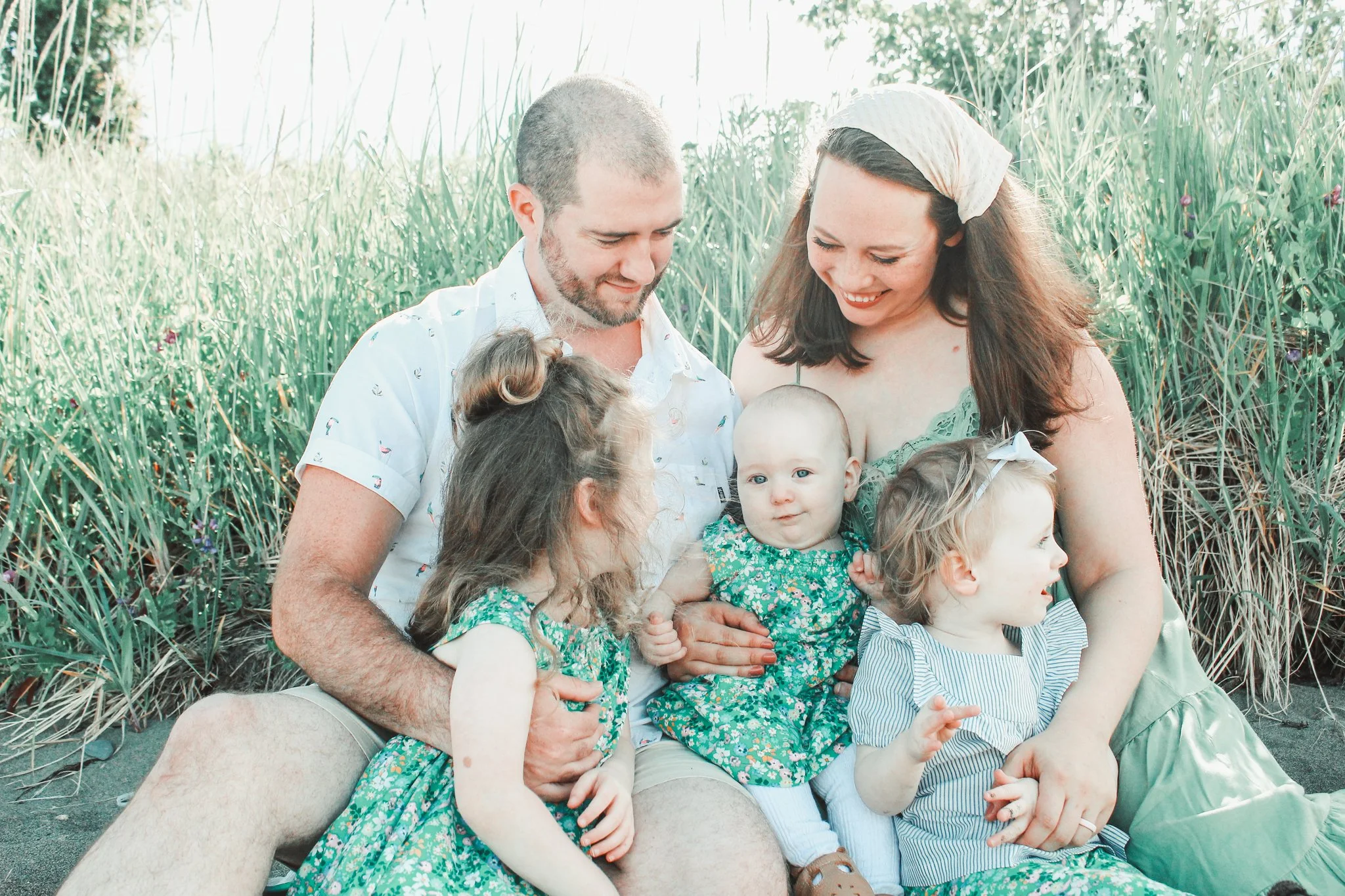 A family of six sitting outdoors among tall grass and flowers, smiling and looking at each other.