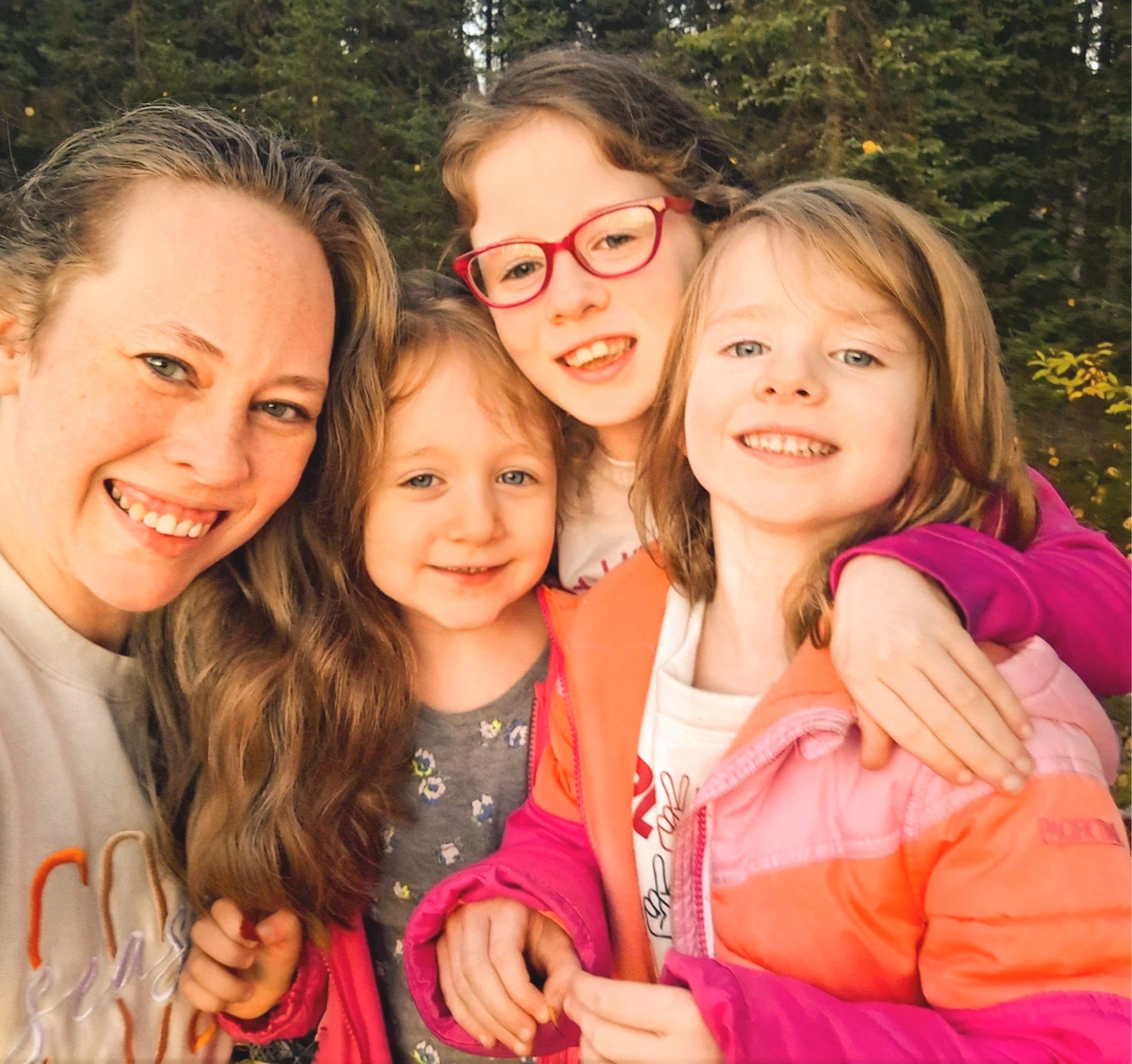 A woman with long, wavy brown hair and four young girls smiling outdoors, with a background of trees.