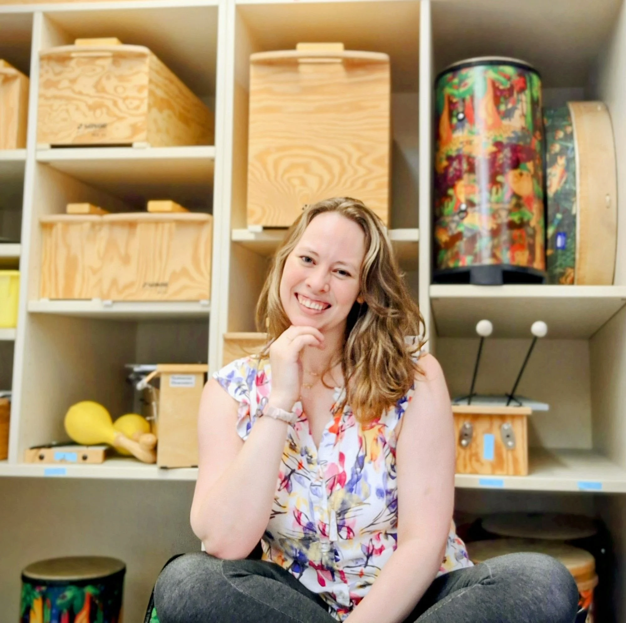 Young woman with wavy blonde hair smiling, posed sitting cross-legged in front of a wall of wooden storage cubbies with colorful objects, wearing a floral dress.