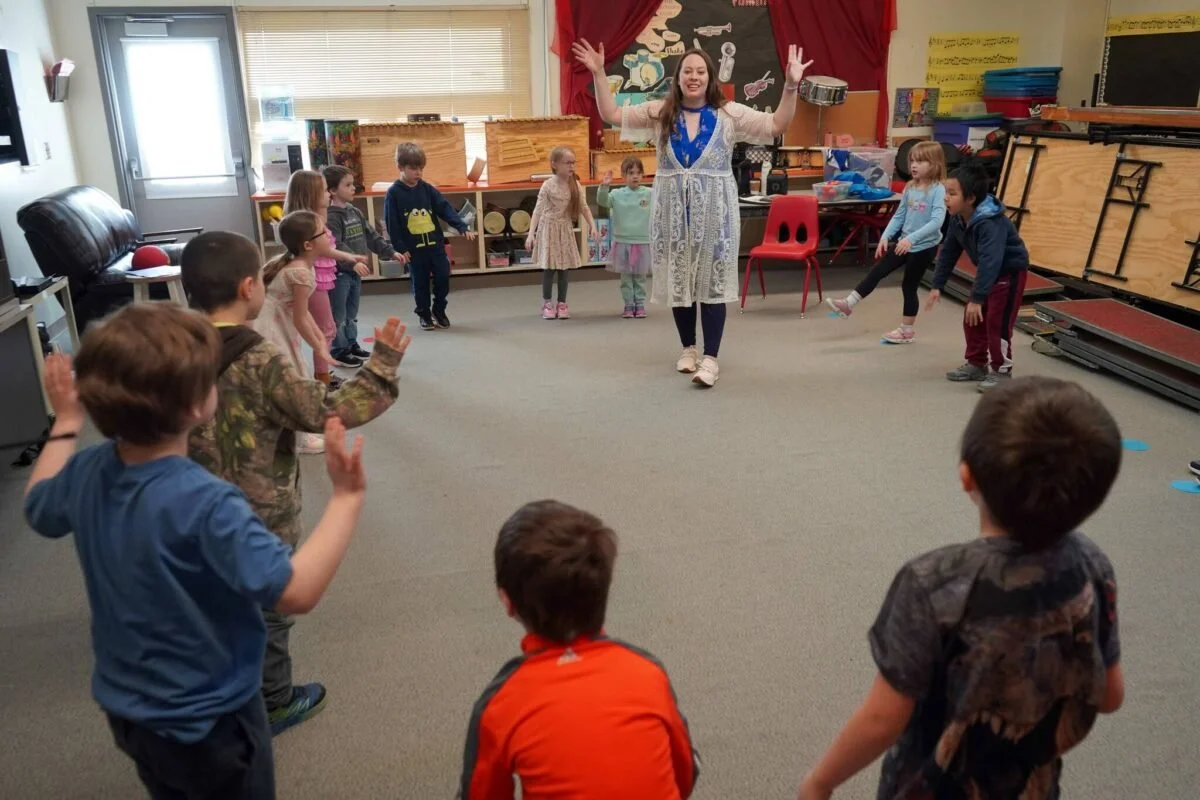 A woman leading a group of young children in an indoor activity, standing with arms raised, in a classroom setting.