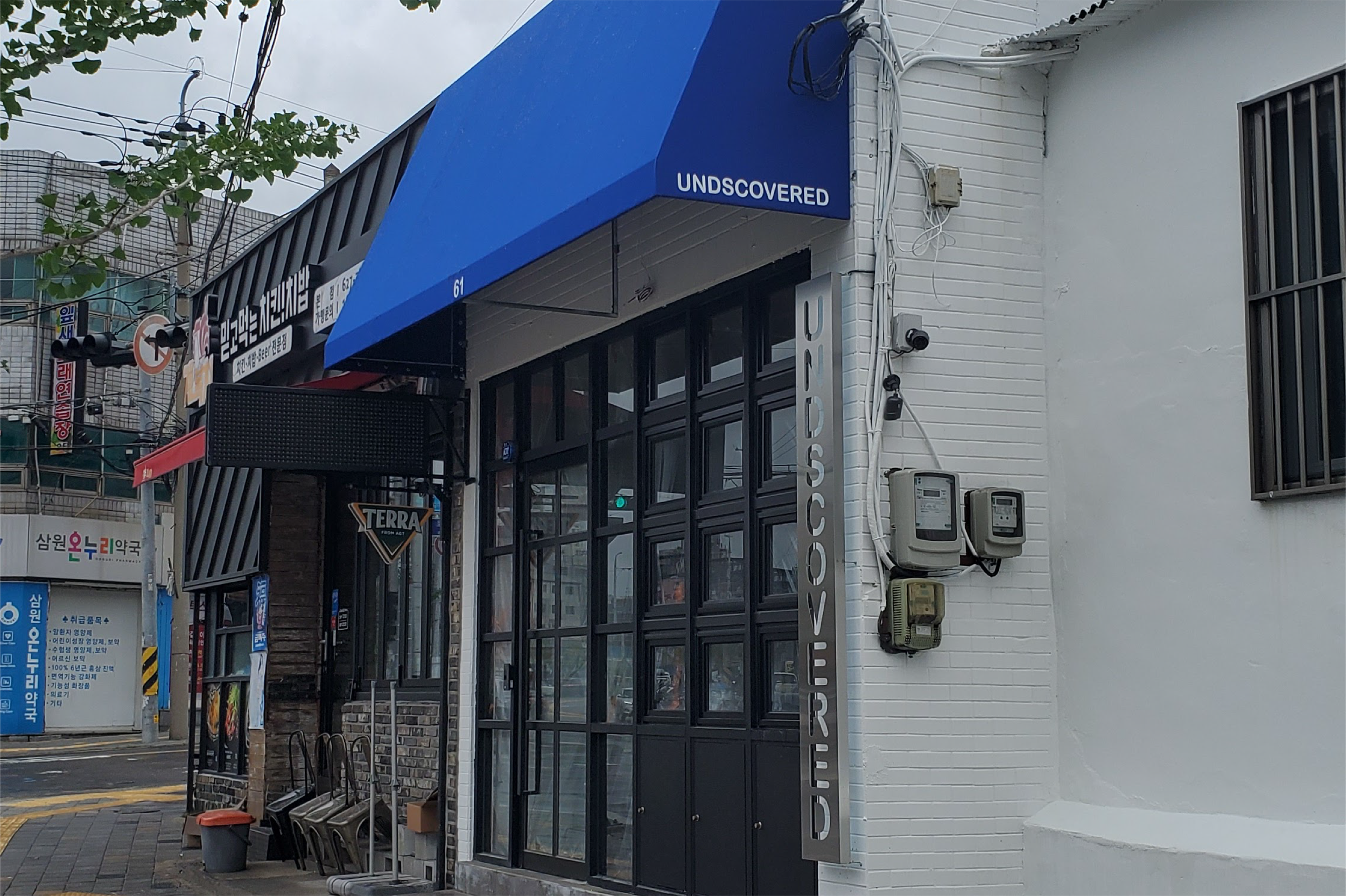 Exterior of a cafe or shop with a blue awning labeled 'UNDCOVERED' and a vertical sign with the same name, black framed glass doors, and two Wi-Fi or electricity meters mounted on the wall. 대구 달서구 성당동 카페, 언디스커버드, 서부정류장 카페, 두류공원 카페