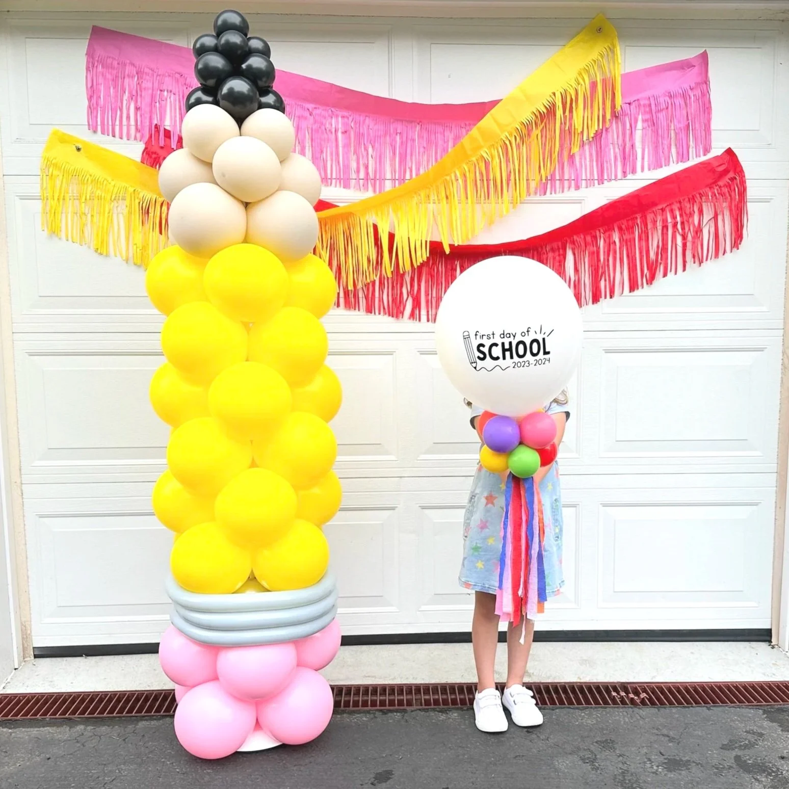 A child standing in front of a decorated garage, holding a large white balloon that reads 'First day of school 2023-2024'. The garage is decorated with colorful fringed banners in pink, yellow, and red. Next to the child is a large balloon arrangement resembling a school bell made of yellow, white, gray, and pink balloons.