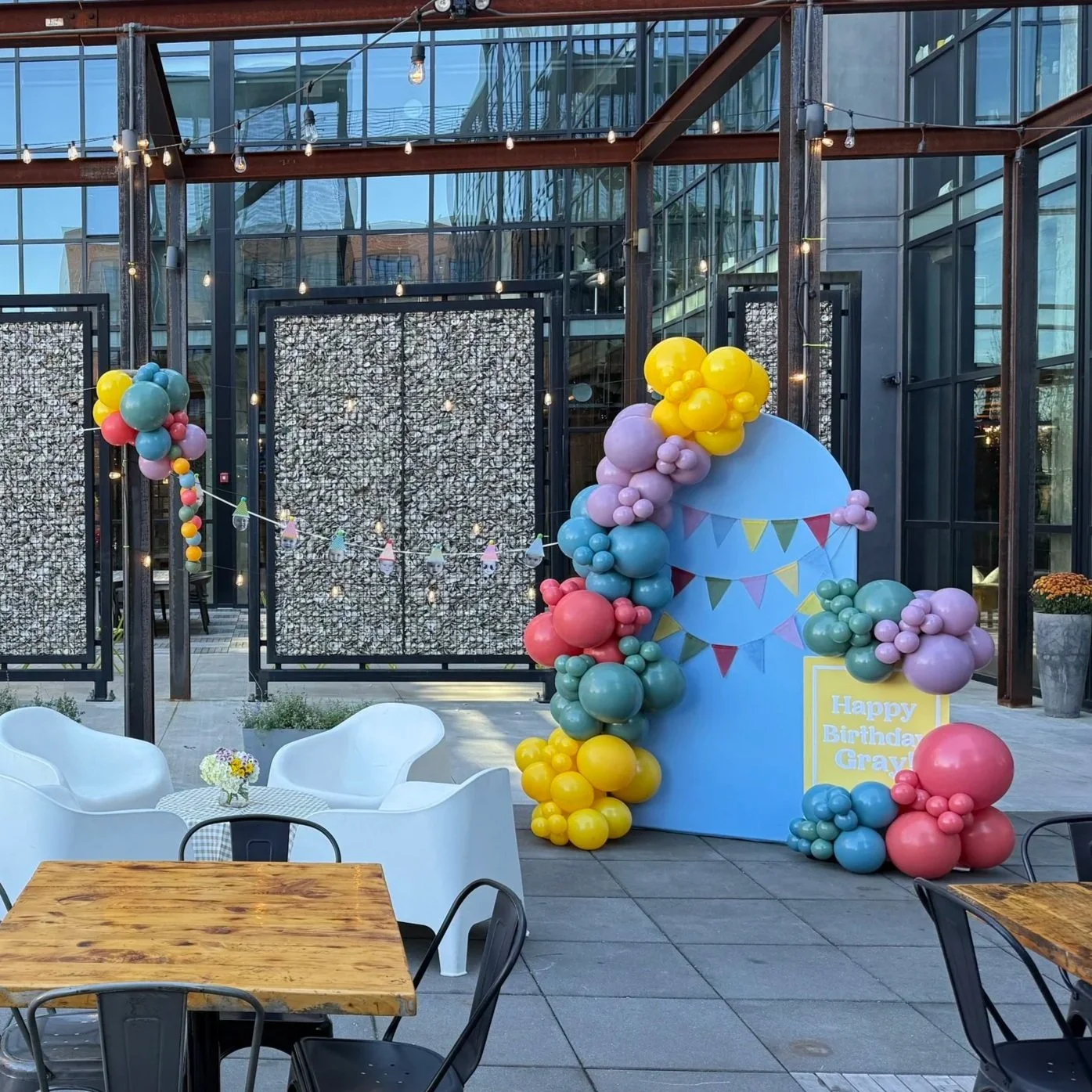 Decorative birthday setup with a large blue cake cutout, colorful balloons, and a sign reading 'Happy Birthday Gray' on an outdoor patio area with tables and chairs, surrounded by glass walls and string lights.