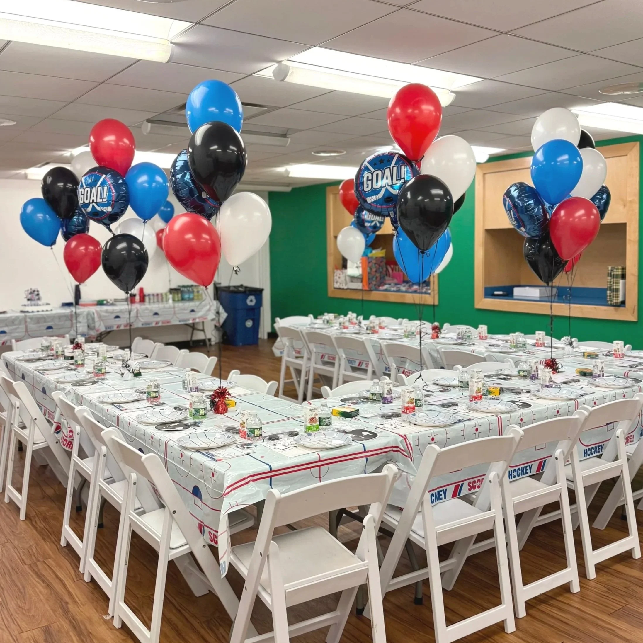 Conference room decorated for a hockey-themed celebration with tables covered in hockey-themed tablecloths, white chairs, and clusters of red, white, blue, black, and hockey-themed balloons.