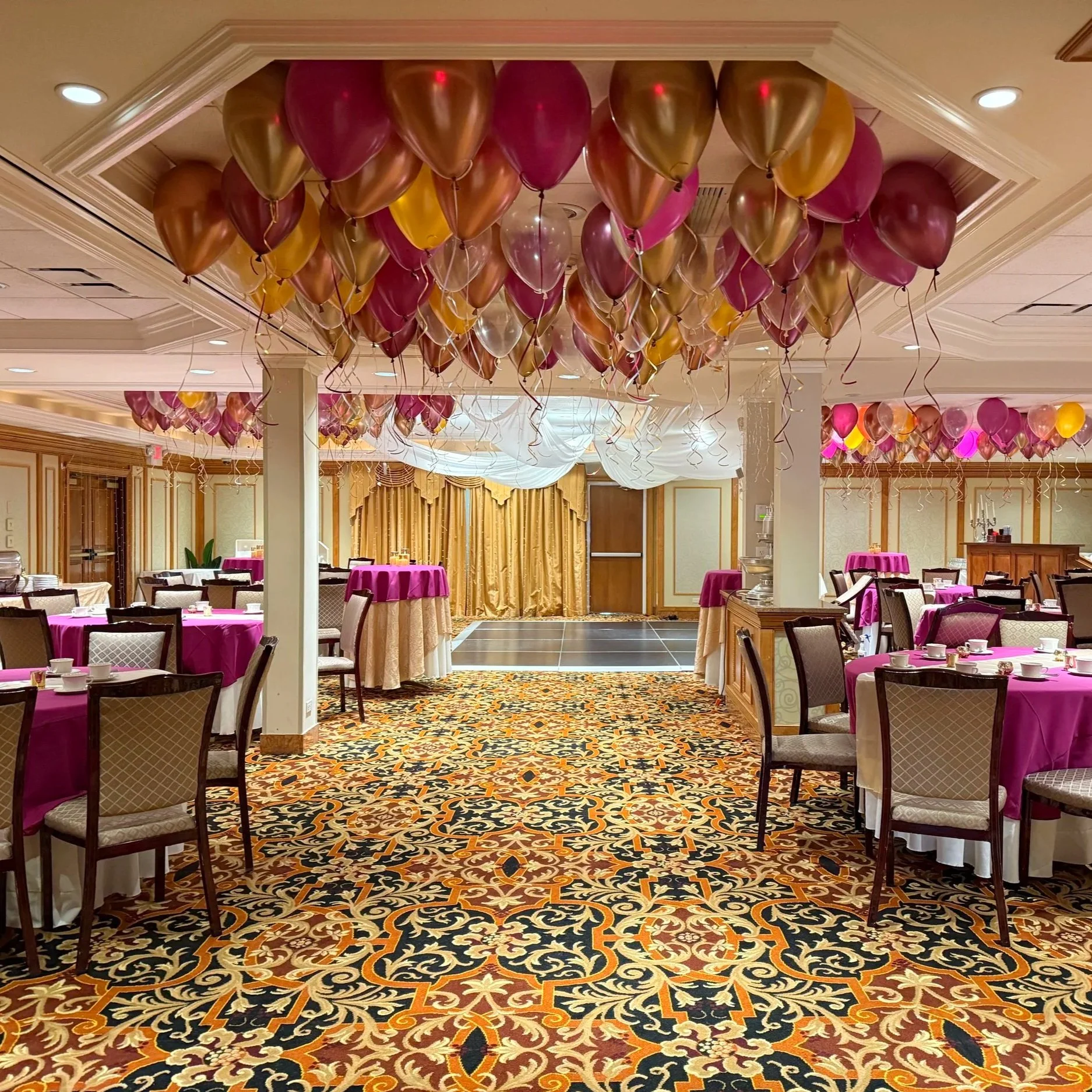A banquet hall decorated for a celebration with pink, gold, and clear balloons hanging from the ceiling. The room features round tables with pink tablecloths and dining settings, beige chairs, and ornate carpet with a floral pattern. Gold curtains and drapes are in the background, with a small stage area.