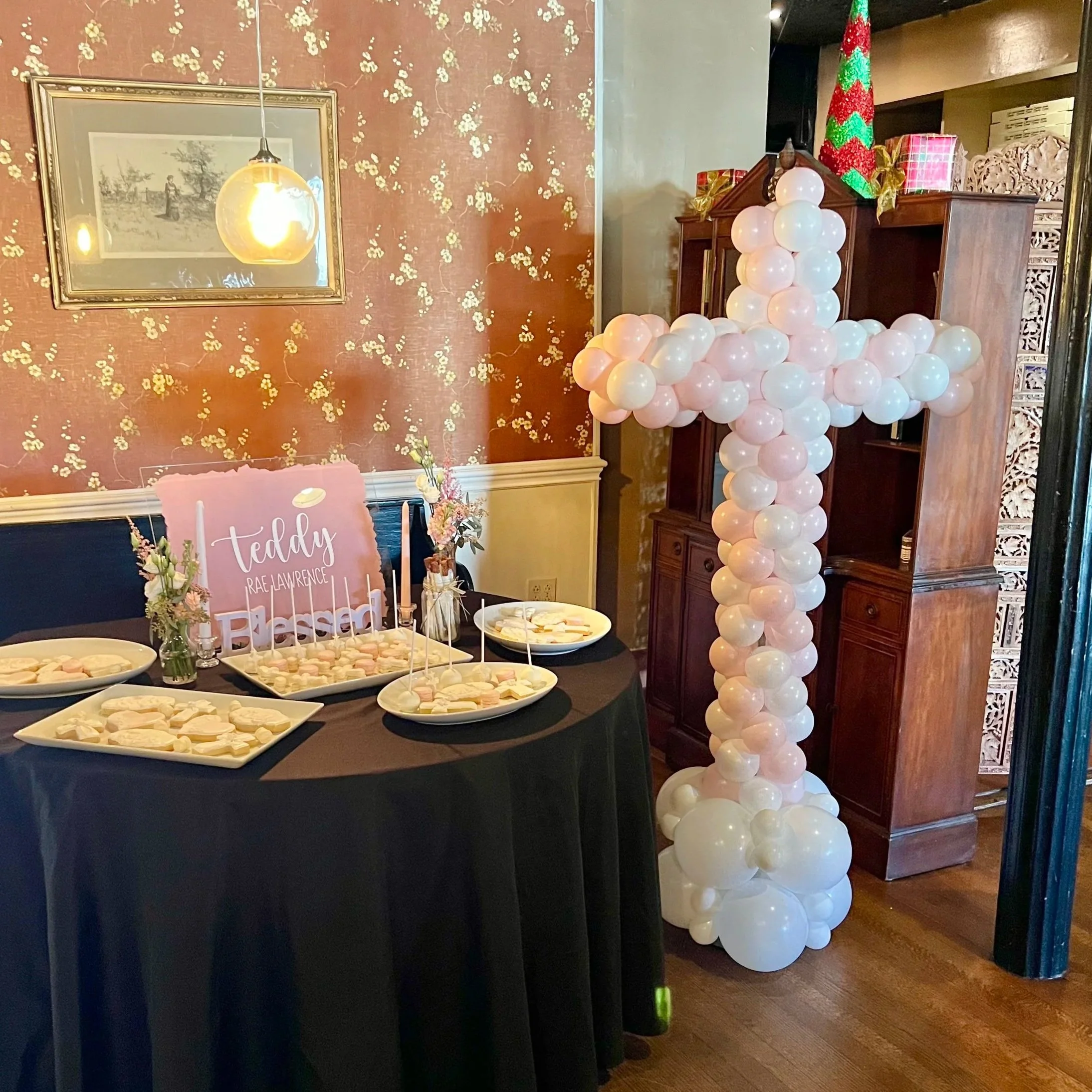 A decor setup for a baby shower with a cross made of white and pink balloons, a table with treats and a pink sign that says 'teddy' and 'Raelynn Lawrence'.
